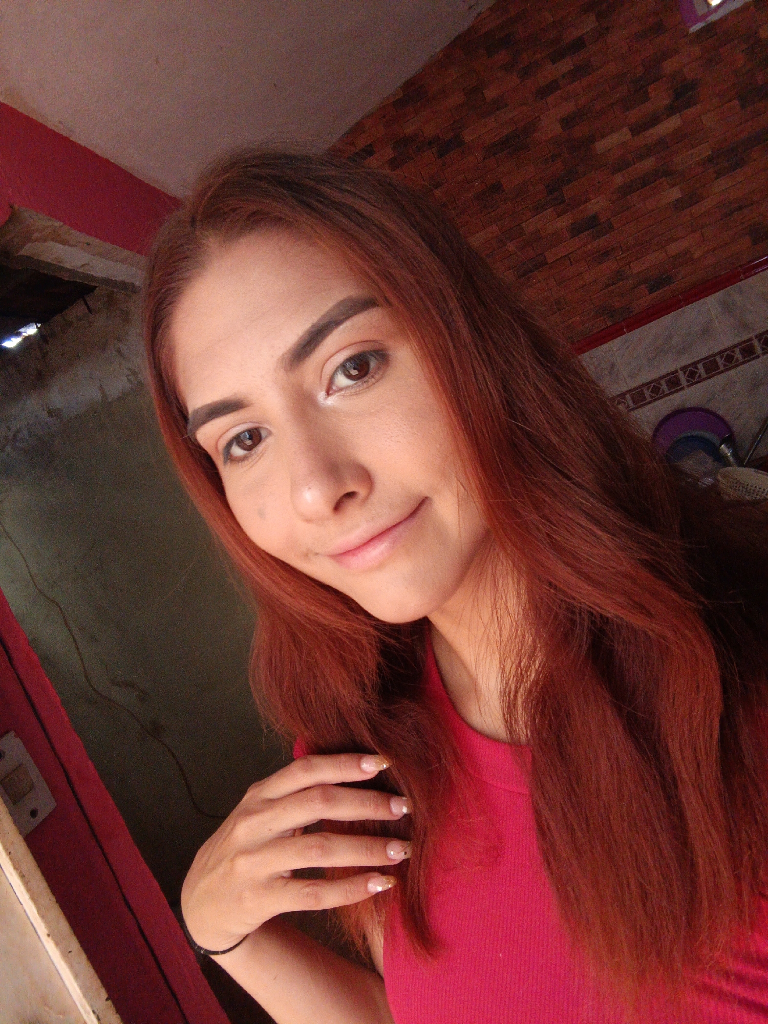 A young adult female with long, auburn hair is pictured in a close-up selfie. She has brown eyes, distinct eyebrows, and a slight smile. She is wearing a red, ribbed tank top. Her left hand, adorned with painted nails, is raised near her chest. A simple black bracelet is visible on her left wrist.
The background is a somewhat cluttered interior space. To the left of the subject is a partly visible door frame painted red and white, with a dark, undefined area beyond it. Behind her is a wall with brown brick tiles. A section of wall beneath the brick tiles is visible, decorated with light-colored tiles with a geometric design. A fan can be seen on the top right corner of the image.
Based on the presence of red-painted elements and the style of architecture in the background, it can be assumed that the location is Juan Griego, Venezuela.