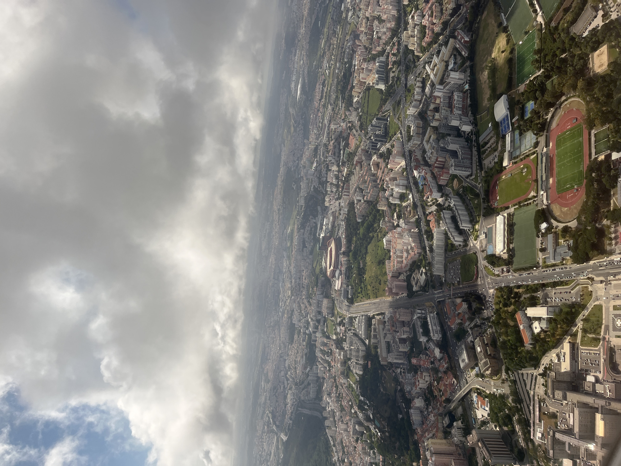 The image shows an aerial view of Lisbon, Portugal, under a cloudy sky. The cityscape is densely populated with buildings and roads. In the foreground, there are sports facilities including a stadium, running track, and athletic fields. In the distance, buildings extend as far as the horizon, partially obscured by clouds and haze. A major road appears to lead towards a large stadium in the middle ground. There are no visible people or animals.