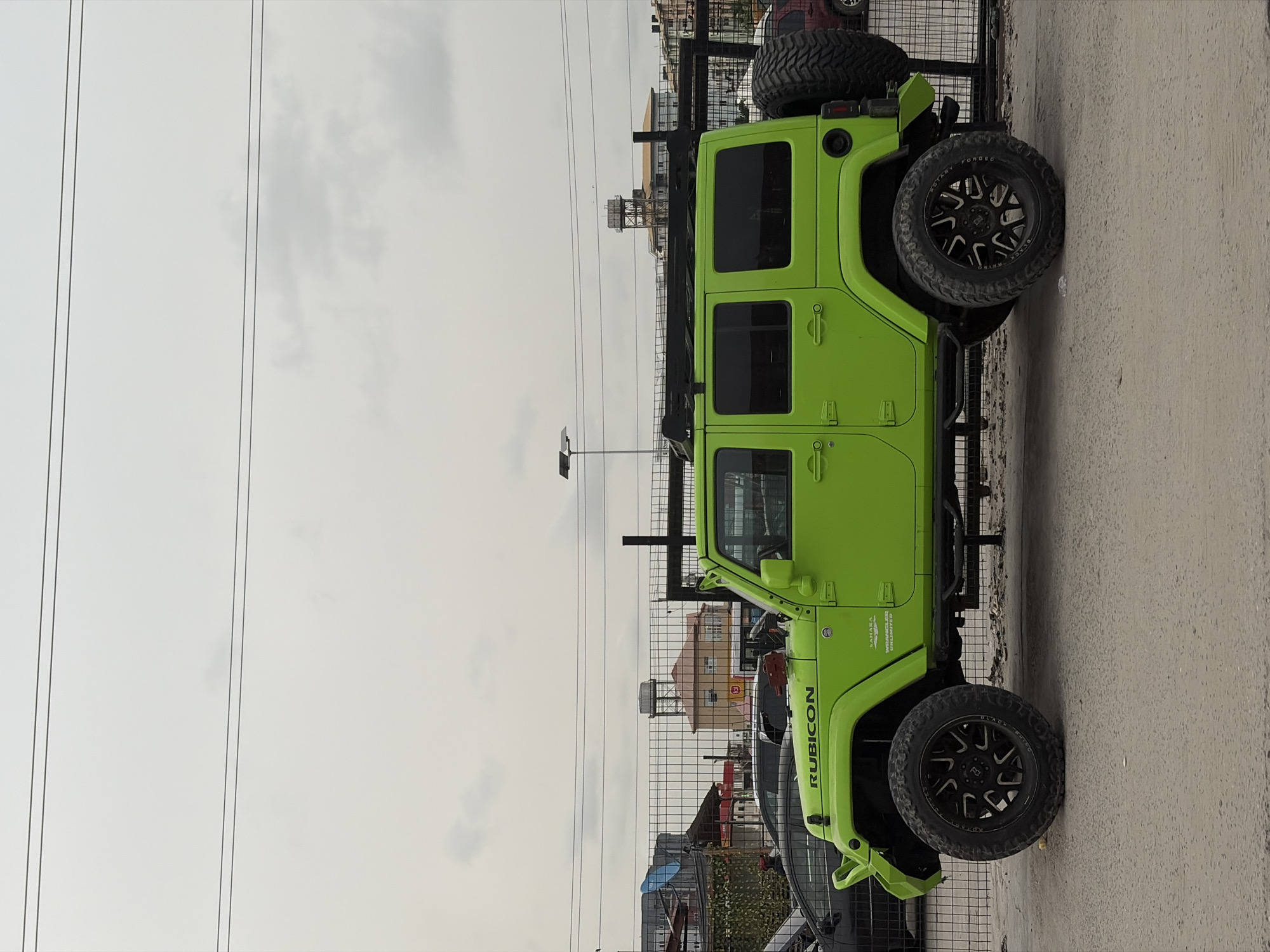 A bright, lime-green four-door Jeep Wrangler Rubicon is prominently featured, parked outdoors with its right side facing the viewer. The vehicle is highly customized, featuring large aftermarket wheels with prominent black rims and rugged tires that show "BLACK RHINO" and "ROTARY FORGED" branding. A spare tire is mounted on the rear. The Jeep also has side steps or rock rails and a roof rack. Text "RUBICON" is clearly visible on the side of the hood, and "WRANGLER UNLIMITED" and "SAHARA" are visible, though partially obscured, on the side panel.
The Jeep is positioned on a light grey concrete or asphalt surface, next to a metal fence. Beyond the fence, several buildings are visible in the background under an overcast, light grey sky with scattered clouds. One building has a water tank on its roof. Overhead, multiple thin power lines stretch across the frame. A street light pole stands to the left of the Jeep. No people are visible in the scene. The setting appears to be an urban or semi-urban area during daytime, possibly in Igboefon, Nigeria.