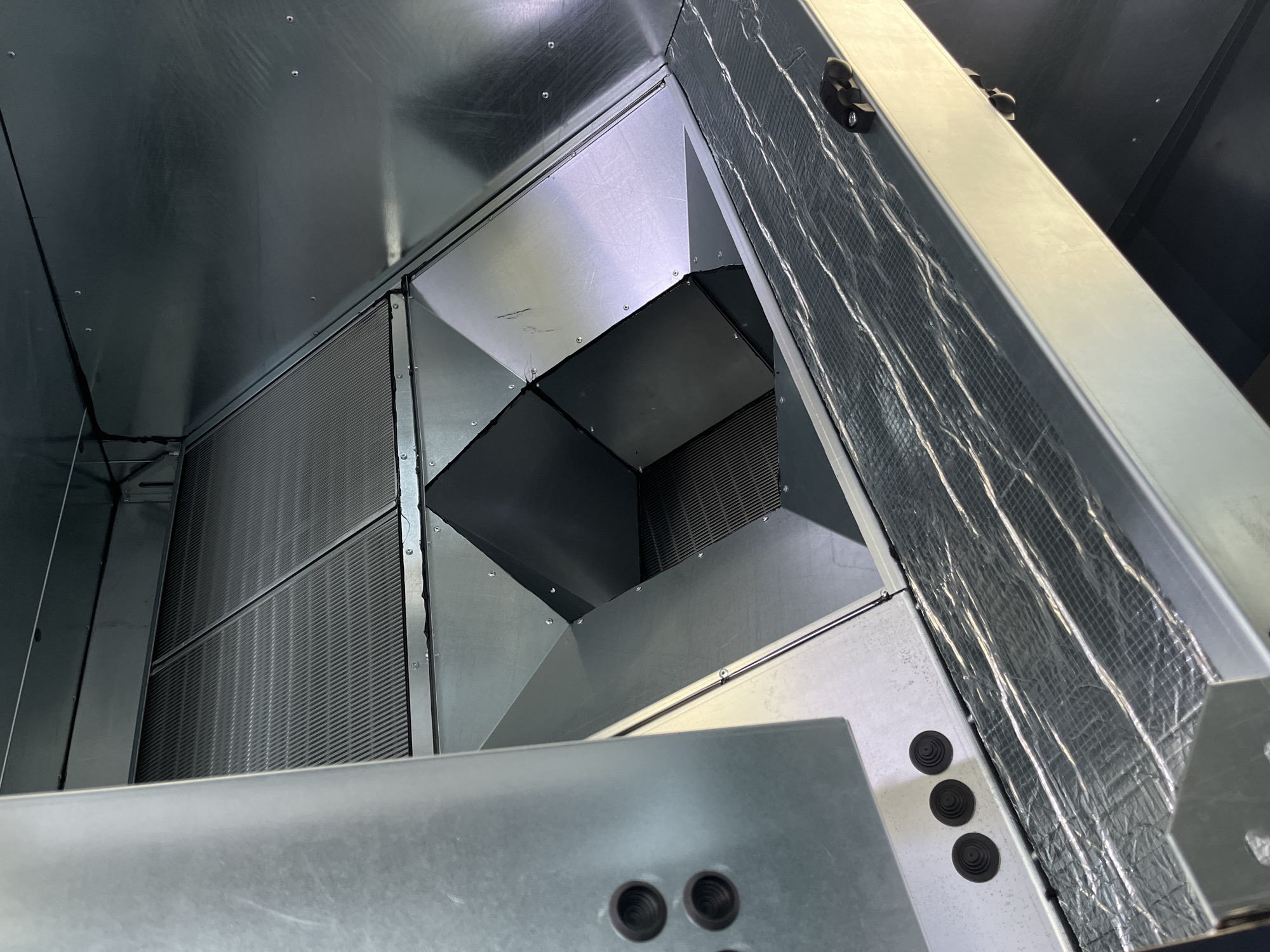 The image shows the interior of an industrial ventilation unit. The unit is constructed from galvanized sheet metal and features a complex, angular internal structure. There are multiple grate-like components visible, likely filters or heat exchangers, and a central, multi-faceted ductwork system. The surfaces are clean and appear to be new or recently maintained. In the foreground, a section of the unit is lined with a silver, textured insulation material. A small black object, possibly a latch or clip, rests on this insulated surface. On the left side of the image, three round, black rubber grommets are visible on a metal panel. The lighting is bright, suggesting an indoor, well-lit environment. There are no people present in the image, and no contextual clues indicate a specific location within Kyiv, Ukraine, beyond the general nature of industrial equipment.