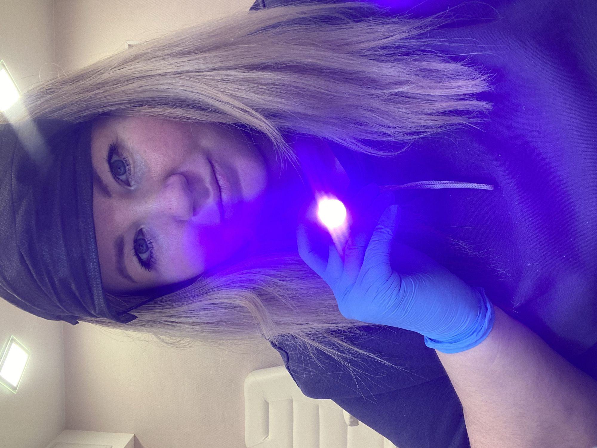 A woman with blonde hair is examining something with a small, bright light held in her gloved hand. She is wearing a dark colored garment and a dark head covering. The environment is a neutral-toned room. It is not possible to verify the location is Chaikovsky, Russia from the given image.