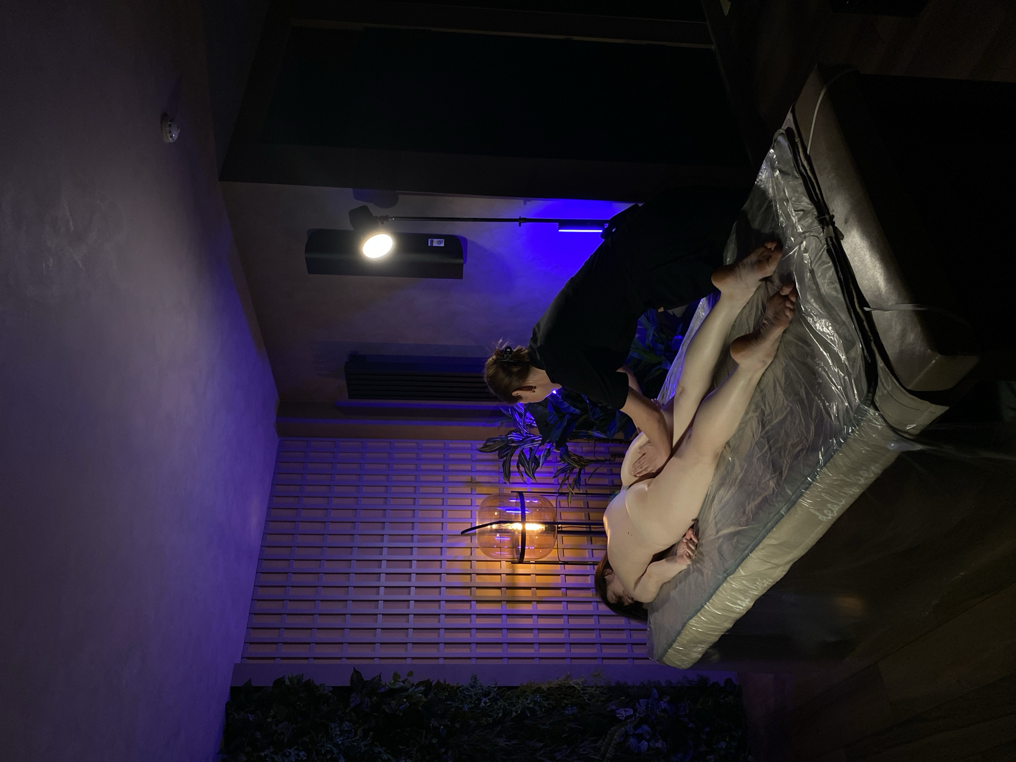 A person in black clothing is performing a massage on a naked person lying on a mattress covered with a plastic sheet. The location appears to be indoors, with a wall featuring a latticed design illuminated by a warm light. There is a plant near the wall. The image is oriented sideways. There is no clear evidence to verify it being in the city of Izhevsk, Russia.