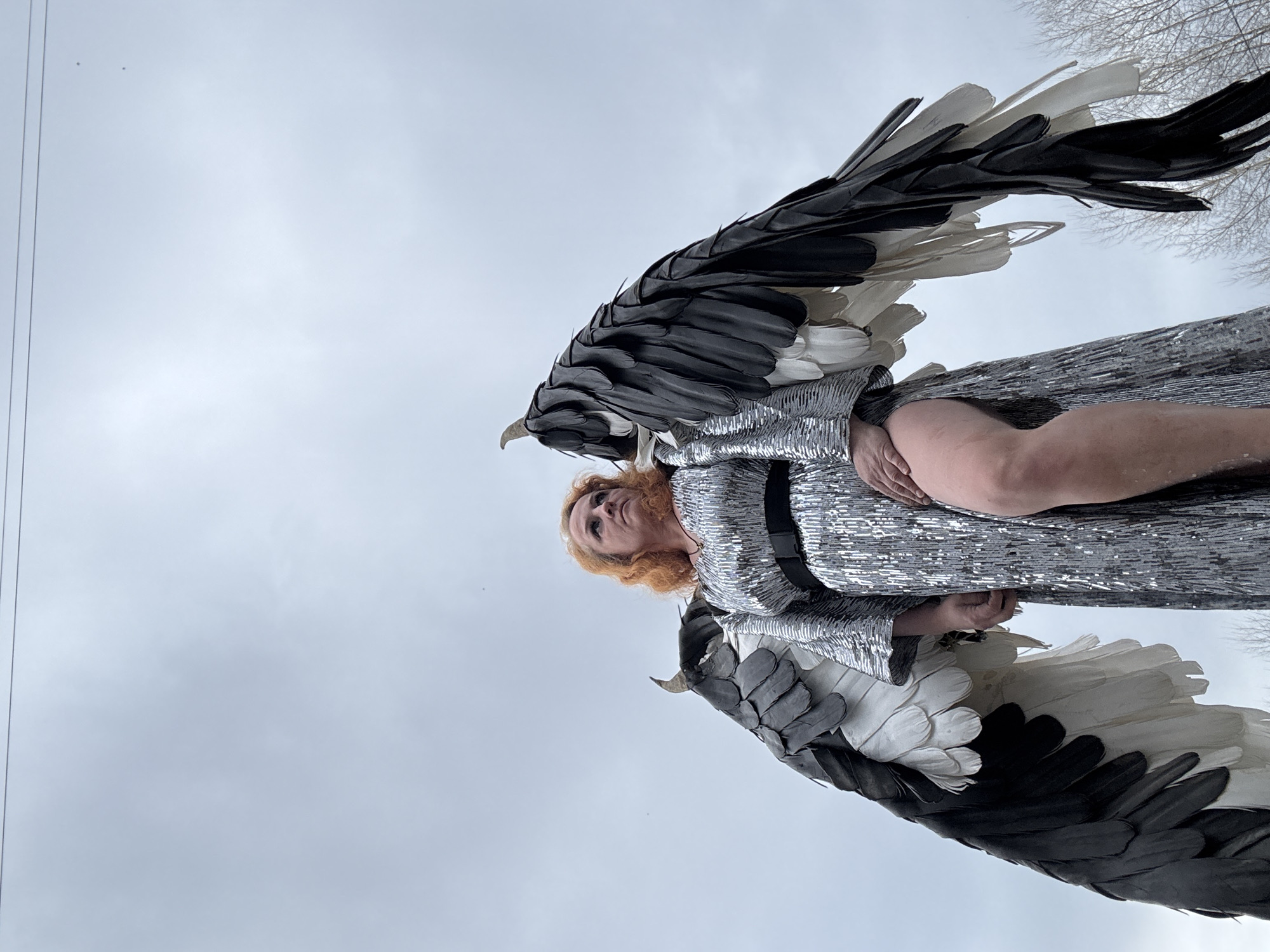 A woman in a silver, sequined dress and large, black-and-white wings is shown in a low-angle shot against a cloudy sky. The woman has red hair and is looking down at the viewer. The background includes bare trees and what appear to be power lines. The location is Chaikovsky, Russia.
