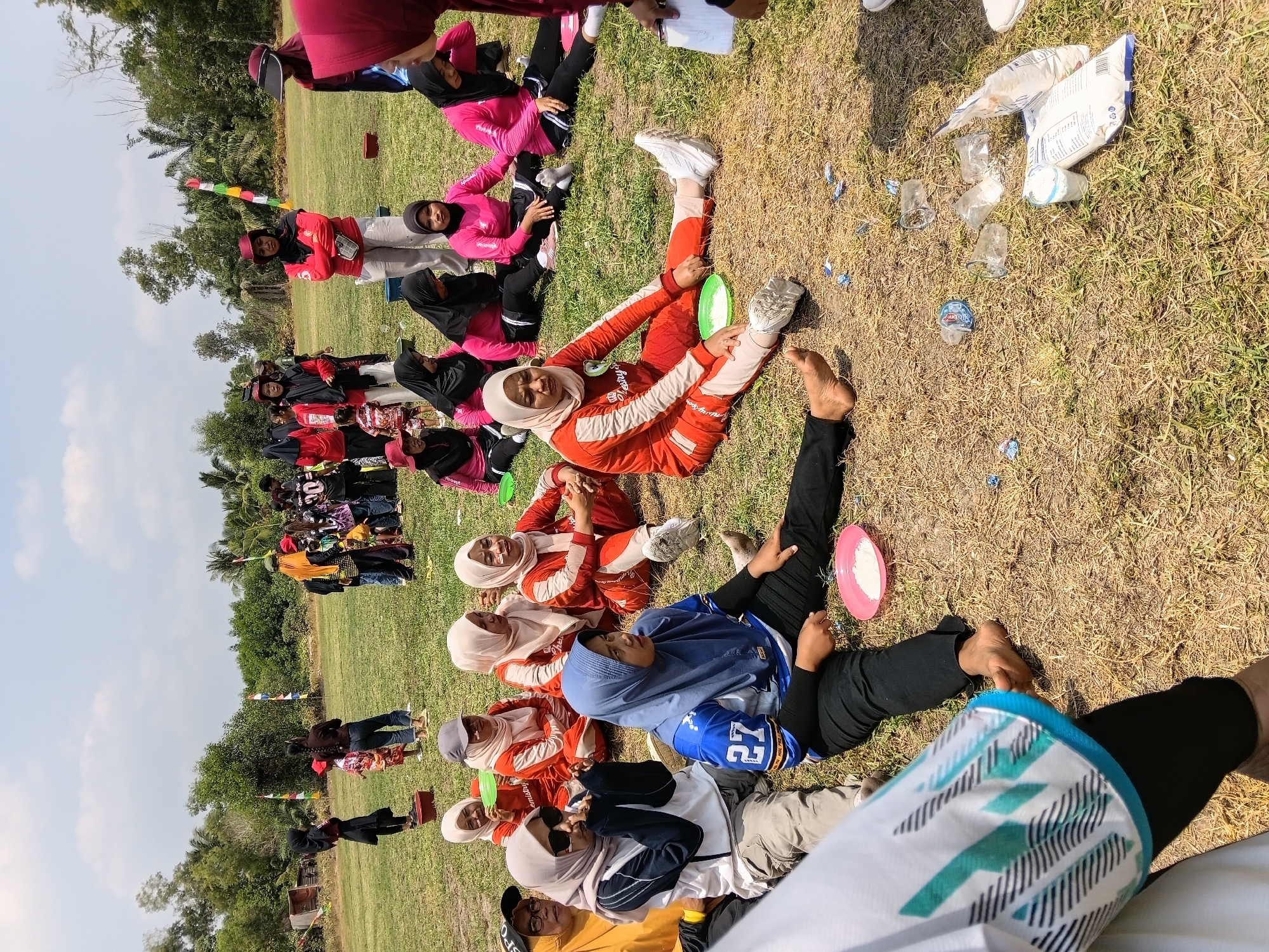 In Margo Rukun, Indonesia, a group of people, predominantly women and girls, are gathered outdoors on a dry, grassy field under a cloudy sky. Many are wearing athletic attire and hijabs. A few individuals are seated on the ground, some with plates of food. Others are standing or in the process of participating in an activity. The scene appears to be a community event or a sporting competition. In the background, trees and foliage are visible, suggesting a rural or semi-rural setting. There are also scattered plastic bottles and food packaging on the ground.