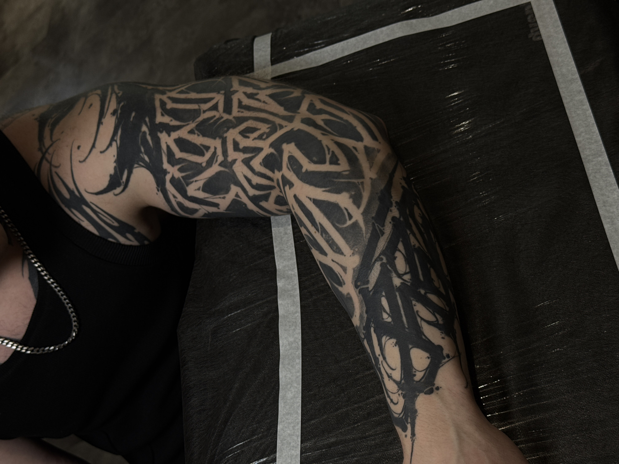 A human arm covered in a large black tattoo is visible. The tattoo consists of abstract geometric shapes, lines, and splatters. The arm extends from the upper left towards the lower right of the frame, resting on a black surface with a light-colored strip running parallel to the arm. A person is wearing a black sleeveless shirt and a silver chain necklace. The surrounding environment is dark. The city and country of Russia and Moscow is not discernible from the image.