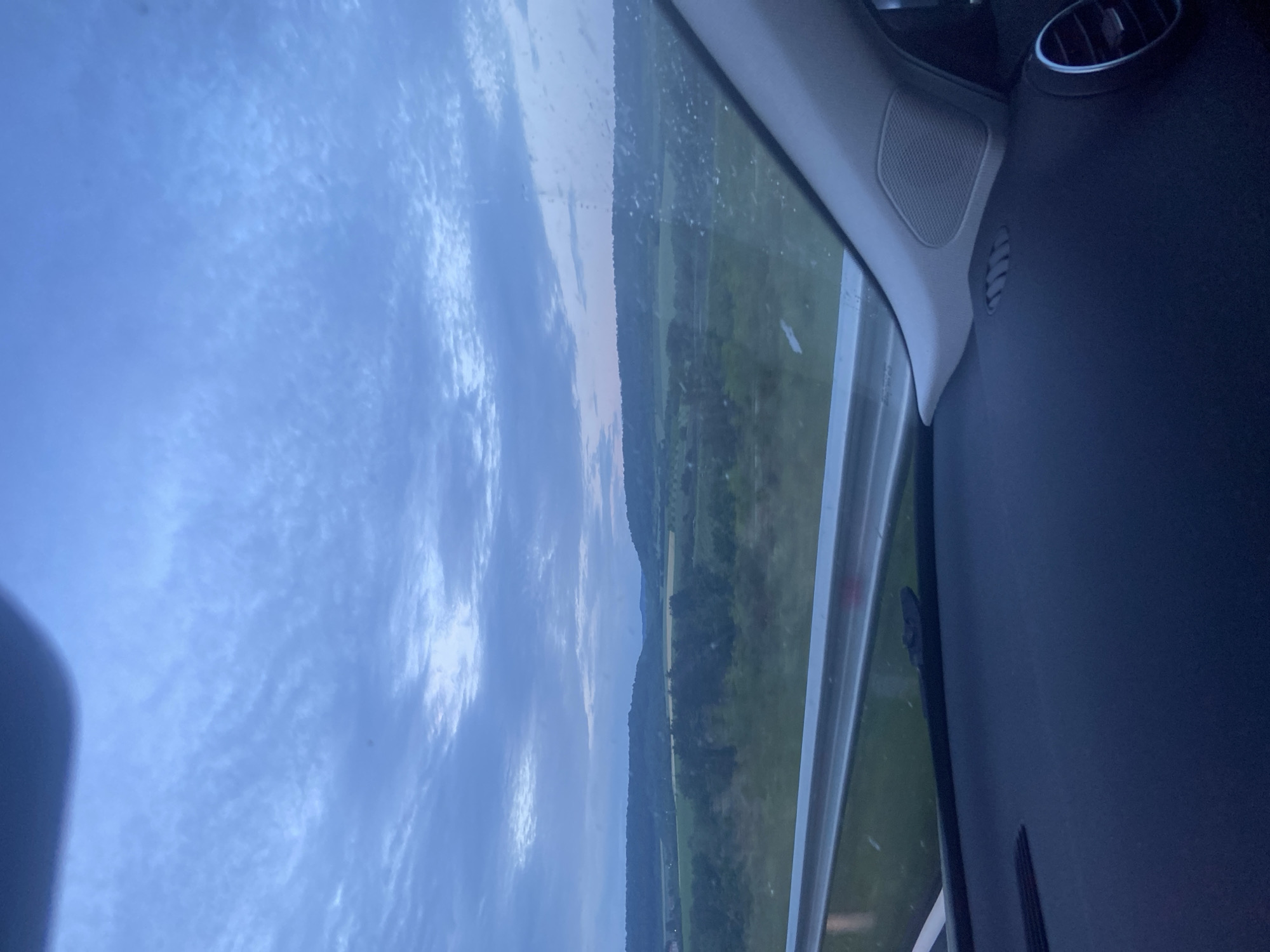 The image captures a serene, rural landscape viewed from the interior of a vehicle. The scene is dominated by a vast sky, a light blue hue filled with scattered white and grey altocumulus clouds, suggesting an overcast or partly cloudy day. Below the sky, the horizon reveals a long stretch of distant, undulating hills or low mountains, covered in dark green vegetation. In the foreground of the exterior view, lush green fields and patches of trees are visible, indicative of agricultural land or natural woodland. The lighting is soft and diffused, contributing to a calm atmosphere, possibly late afternoon or early evening.
On the right side, the interior of a modern car is visible. Key elements include a light grey A-pillar, which houses a circular speaker grille. Adjacent to this, a section of a dark dashboard is seen, featuring a circular air vent with metallic trim. The window frame and sill are also discernible, framing the exterior view. The composition implies a journey through a scenic area. No people are visible, nor is any discernible text present in the image. The general landscape is consistent with a rural setting in a region like Dragijevo, Bulgaria.