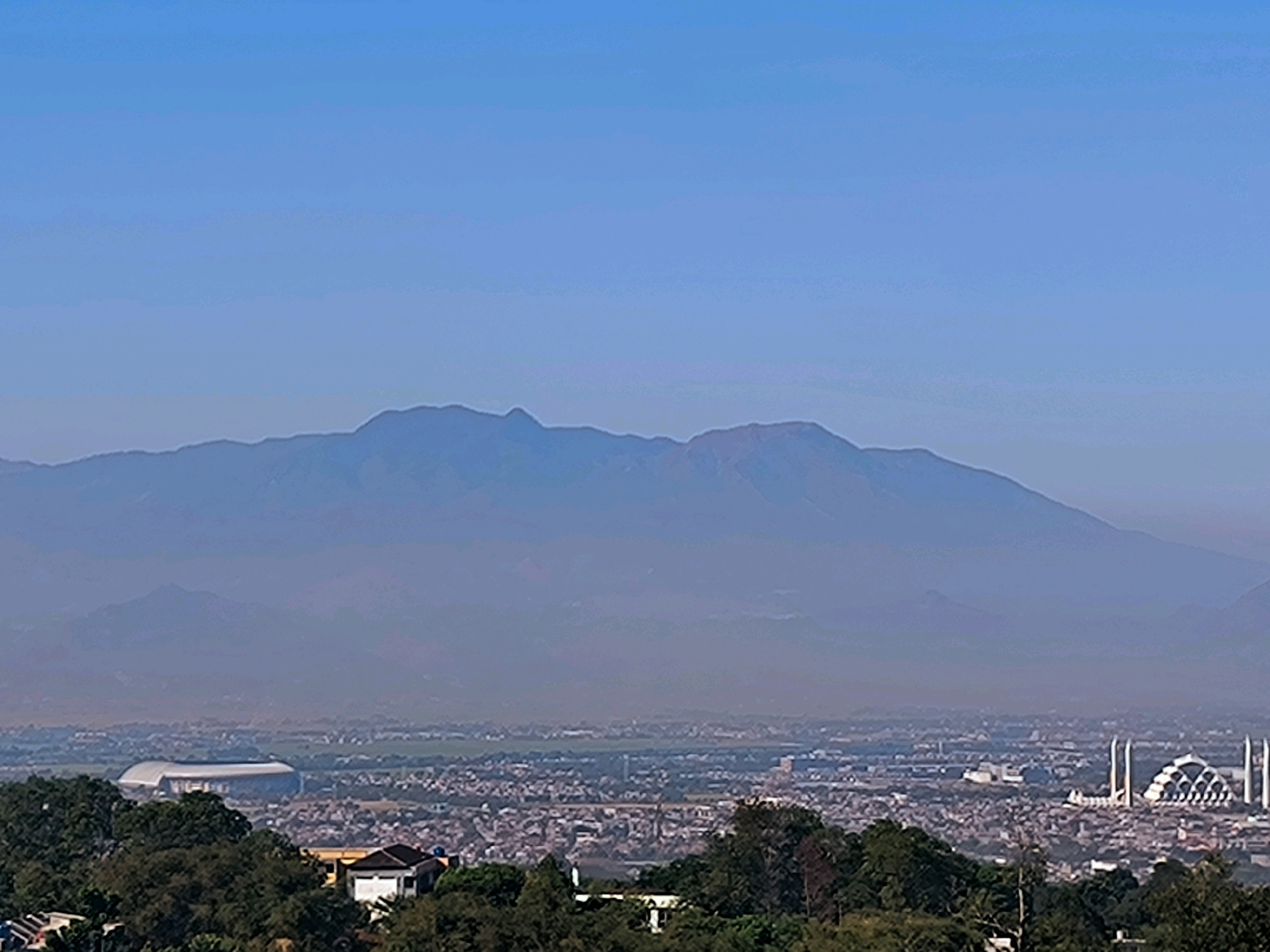 This image depicts a panoramic view of Bandung City, Indonesia, dominated by a hazy mountain range in the background. In the foreground, lush green trees and some houses are visible, suggesting an elevated viewpoint. Mid-ground features a sprawling urban landscape with a stadium on the left and a distinctive mosque with multiple minarets on the right. The city appears densely populated with buildings. The overall atmosphere is serene, with a clear blue sky above.