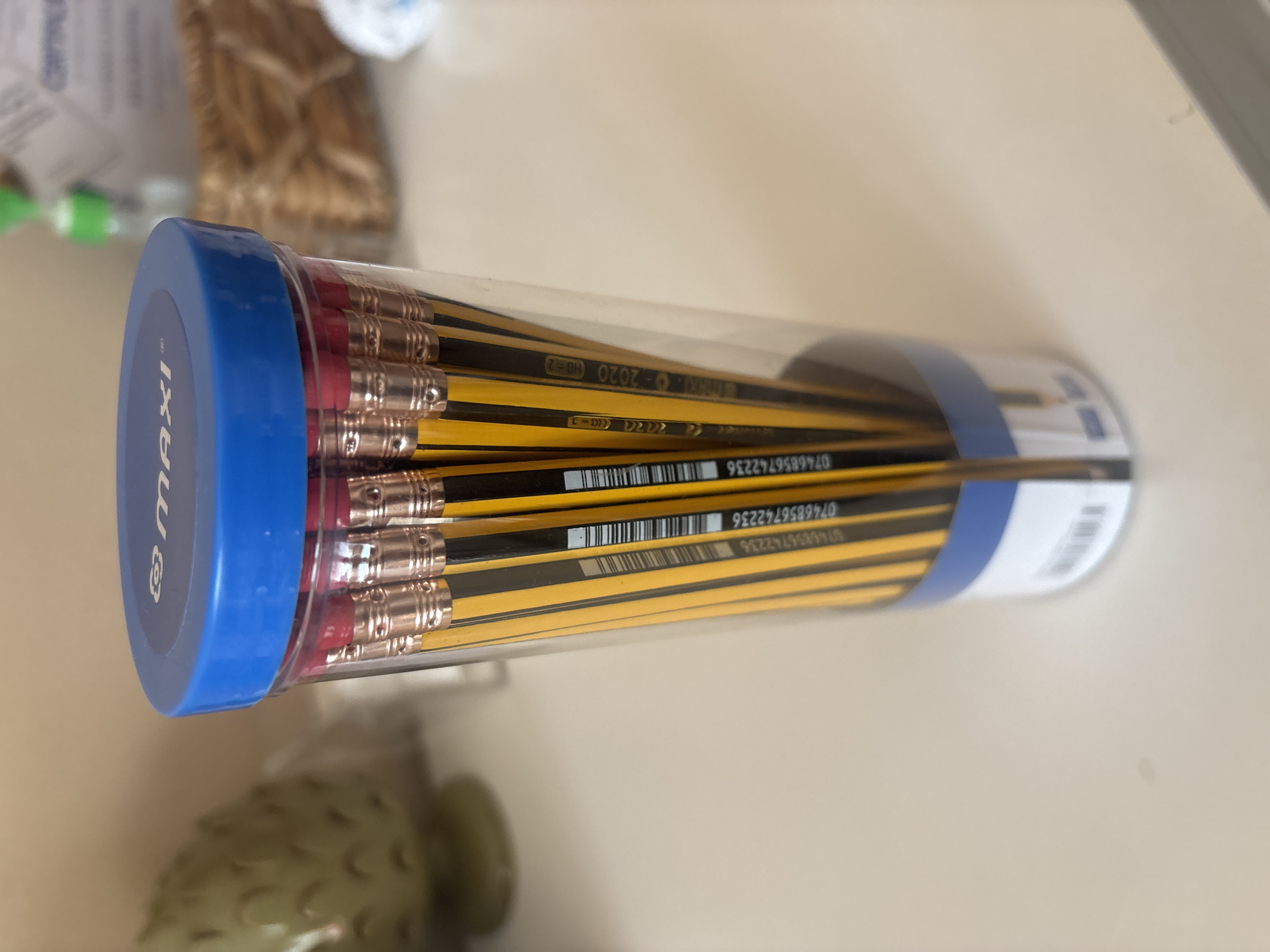 A transparent cylindrical container holds a bundle of yellow and black striped pencils. The container has a blue lid with the word "maxi" imprinted on it in white. The pencils are all topped with pink erasers and metal ferrules. Several pencils have visible barcodes and text printed on their barrels, including what appears to be "0746856742236" and "HB-2 2020". The container and pencils are on a light-colored, possibly wooden surface. In the background, out of focus, is a light green, textured decorative object and a woven basket. The overall scene is indoors, likely on a desk or table. There is no indication of the location being Dubai, United Arab Emirates.