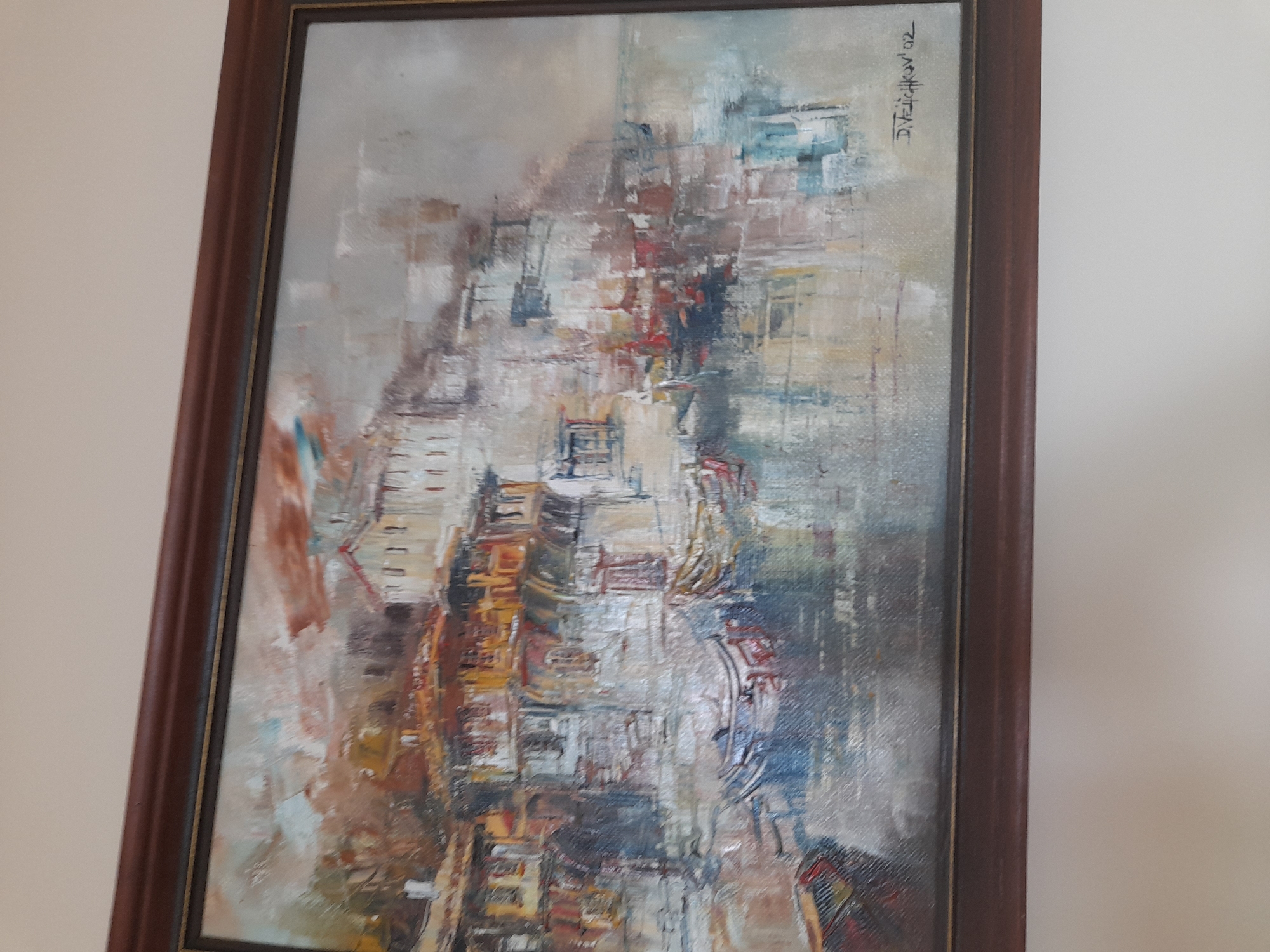 The image shows a framed abstract painting. The painting depicts an impressionistic cityscape with buildings rendered in shades of gray, white, brown, yellow, red, and blue. The brushstrokes are visible and create a sense of movement and texture. The painting is signed "D. Velichkov '02" in the upper right corner. The frame is dark brown. The painting is hanging on a light-colored wall. Without more information, it is impossible to determine if the cityscape is meant to be Sofia, Bulgaria.