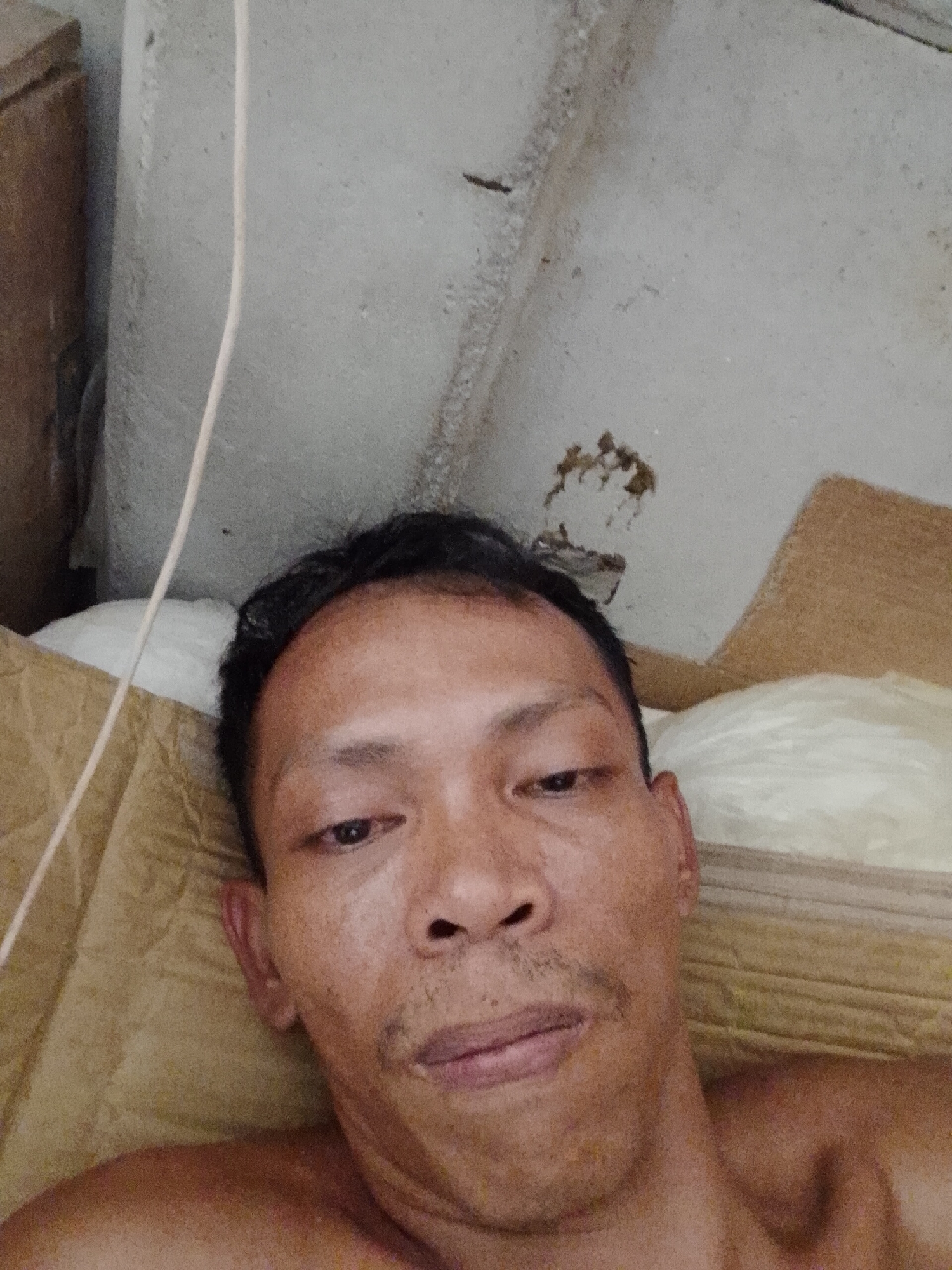 A selfie of an Asian man with dark hair, lying down. He has light-brown skin and stubble on his chin and upper lip. He is positioned against a backdrop of what appears to be a concrete wall with some discolored spots and a white cable running along it. Beside the wall, is brown cardboard and a white pillow. The photograph was taken in an interior location, probably in Bukit Ketok, Indonesia, but specific details about the environment are limited to the immediate vicinity of the subject.