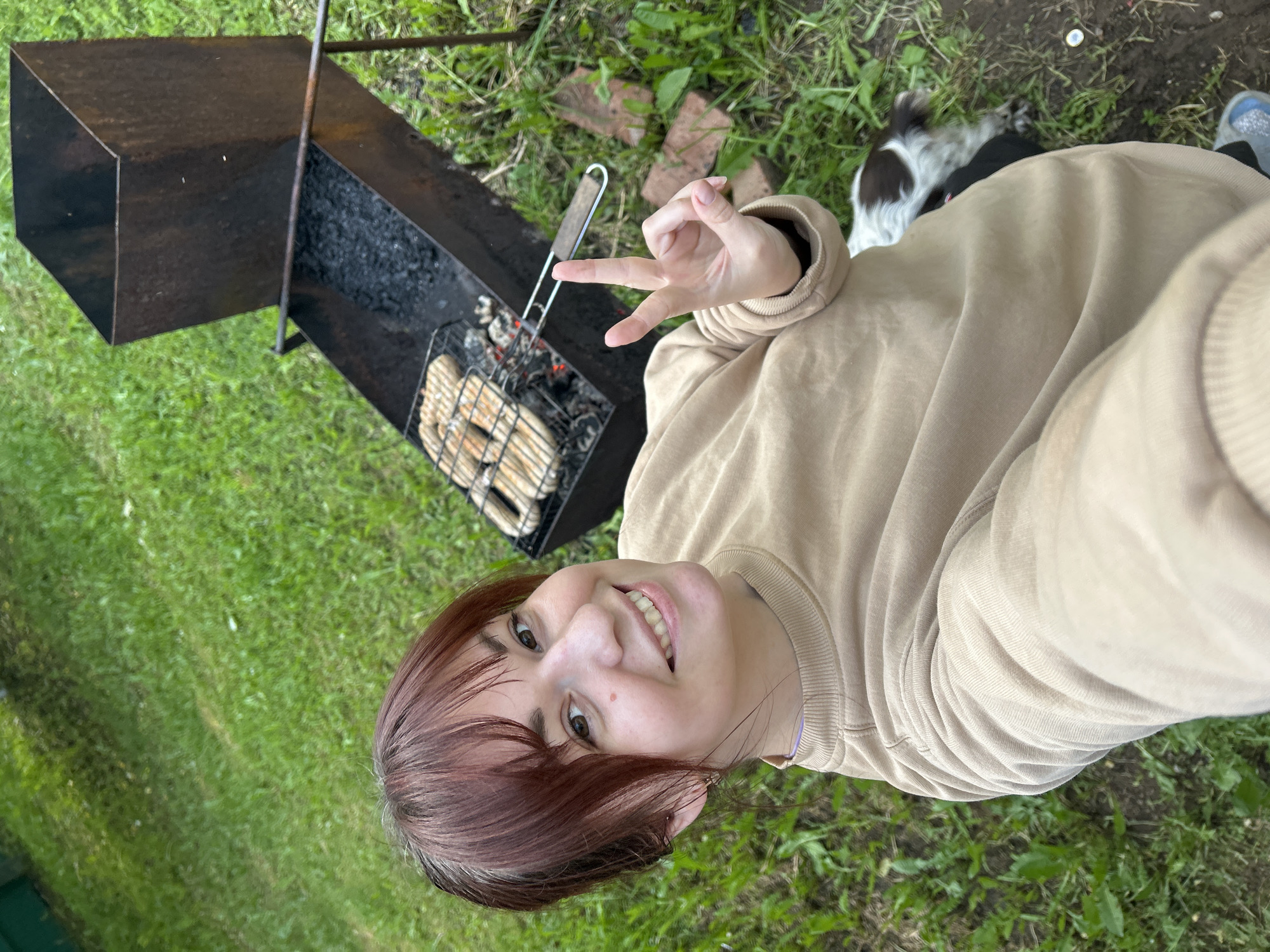 A woman with auburn hair wearing a beige sweatshirt is standing on a green lawn next to a metal barbecue grill. The grill contains coals and food in a wire basket. The woman is giving a peace sign with her hand. Part of a dog can be seen behind her. Some bricks are on the ground near the grill. The location is Фоки, Russia.
