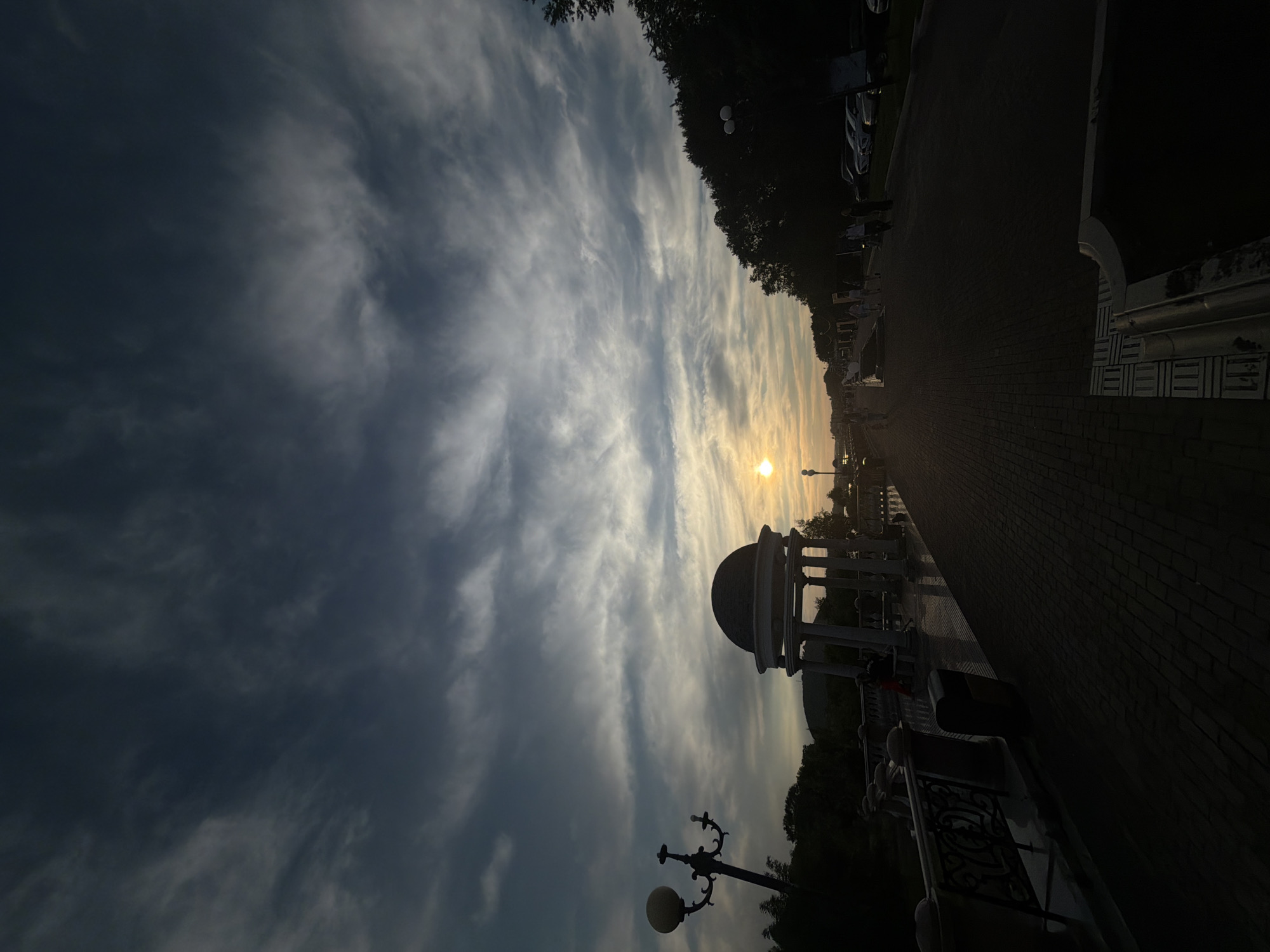 The image depicts a sunset scene on a promenade in Birobidzhan, Russia. A distinctive white rotunda with a domed roof stands prominently on the left side of the promenade. A tall, ornate lamppost is also visible near the rotunda. The promenade itself is paved with brickwork and is lined with a decorative railing. Several individuals are walking along the promenade in the distance, and cars are parked on the right side of the scene. The sky is filled with dramatic, wispy clouds, and the setting sun is visible as a bright orb near the horizon. Trees and other foliage form a dark silhouette against the illuminated sky on the far right. The lighting suggests it is late afternoon or early evening.