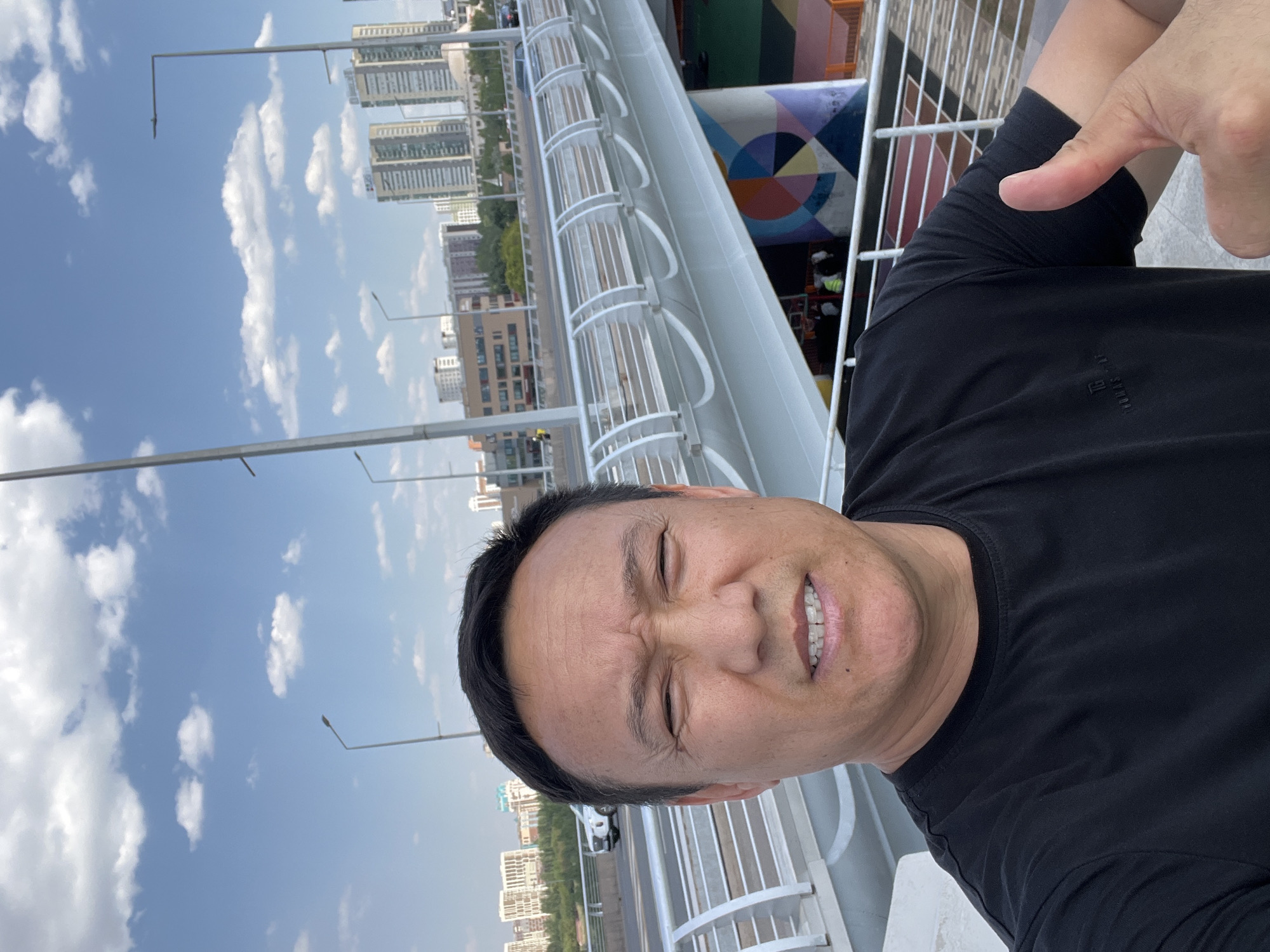 A man takes a selfie on a bright, sunny day in Astana, Kazakhstan, on a pedestrian walkway of a modern overpass. He is wearing a black t-shirt and gives a thumbs-up with his right hand. The overpass features white, curved railings and streetlights. In the background, the cityscape of Astana is visible, with numerous tall buildings under a blue sky with scattered white clouds. The lower portion of the overpass is decorated with a large, colorful geometric mural. A white car is visible driving on the road beneath the overpass.