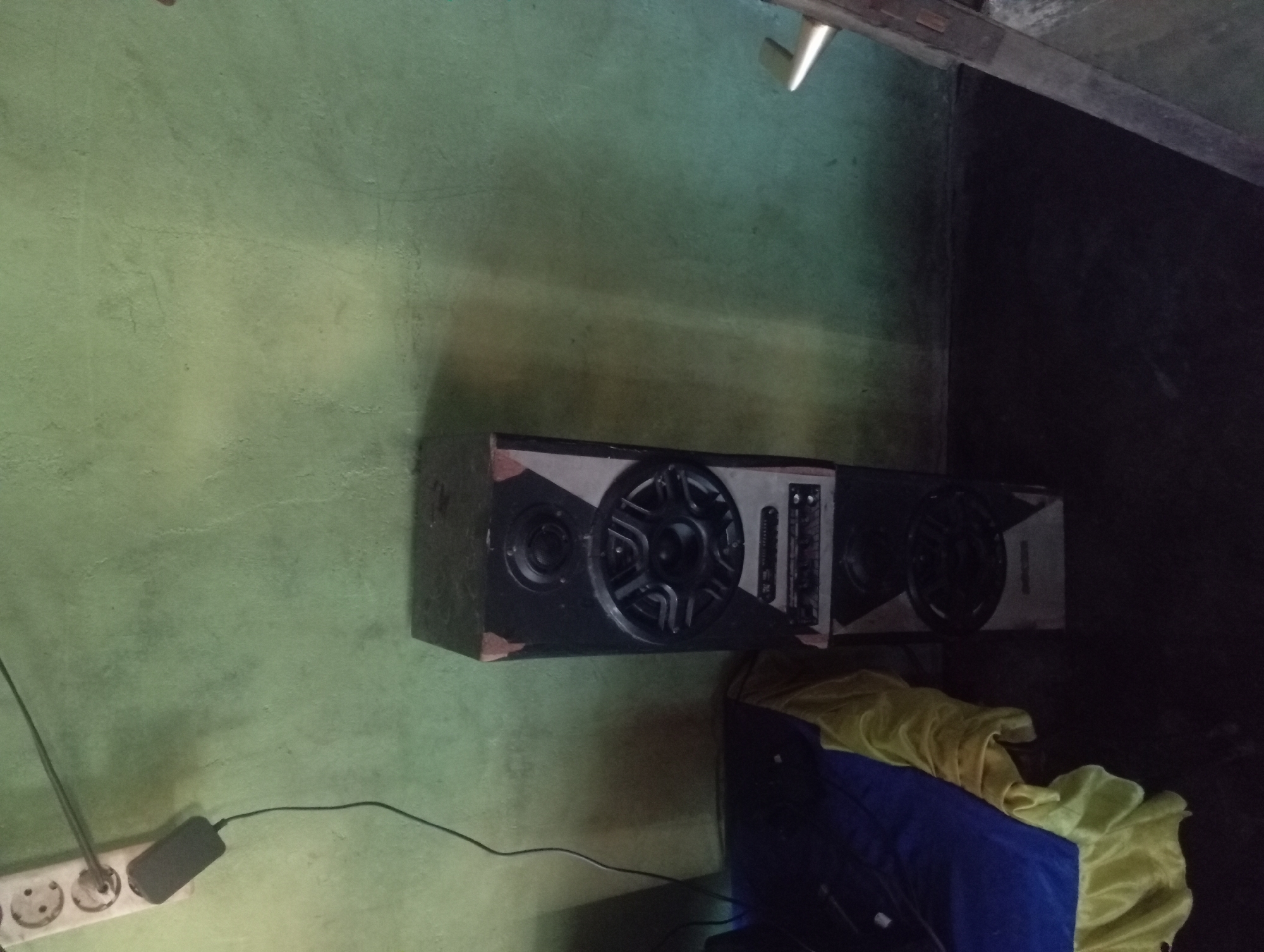 The image depicts a corner of a room with green walls and a dark floor. In the center of the frame, two stacked speakers are visible. The top speaker is a tall, rectangular unit with a black face, featuring a tweeter at the top and a larger mid-range speaker below. Below the mid-range speaker, there's a silver panel with various controls and buttons. The bottom speaker is also rectangular and black, with a large speaker grille. To the left of the speakers, a blue and yellow cloth is draped over an unseen object, partially obscuring the lower part of the wall. In the upper left corner, a white electrical outlet strip is mounted on the wall, with a black power adapter plugged into one of the sockets and a thin black wire extending downwards. A hint of a wooden structure, possibly a door frame or furniture, is visible on the right side of the image. The lighting is dim, casting shadows on the floor and walls. The scene appears to be inside a residential or possibly a workshop area in Rejosari, Indonesia.