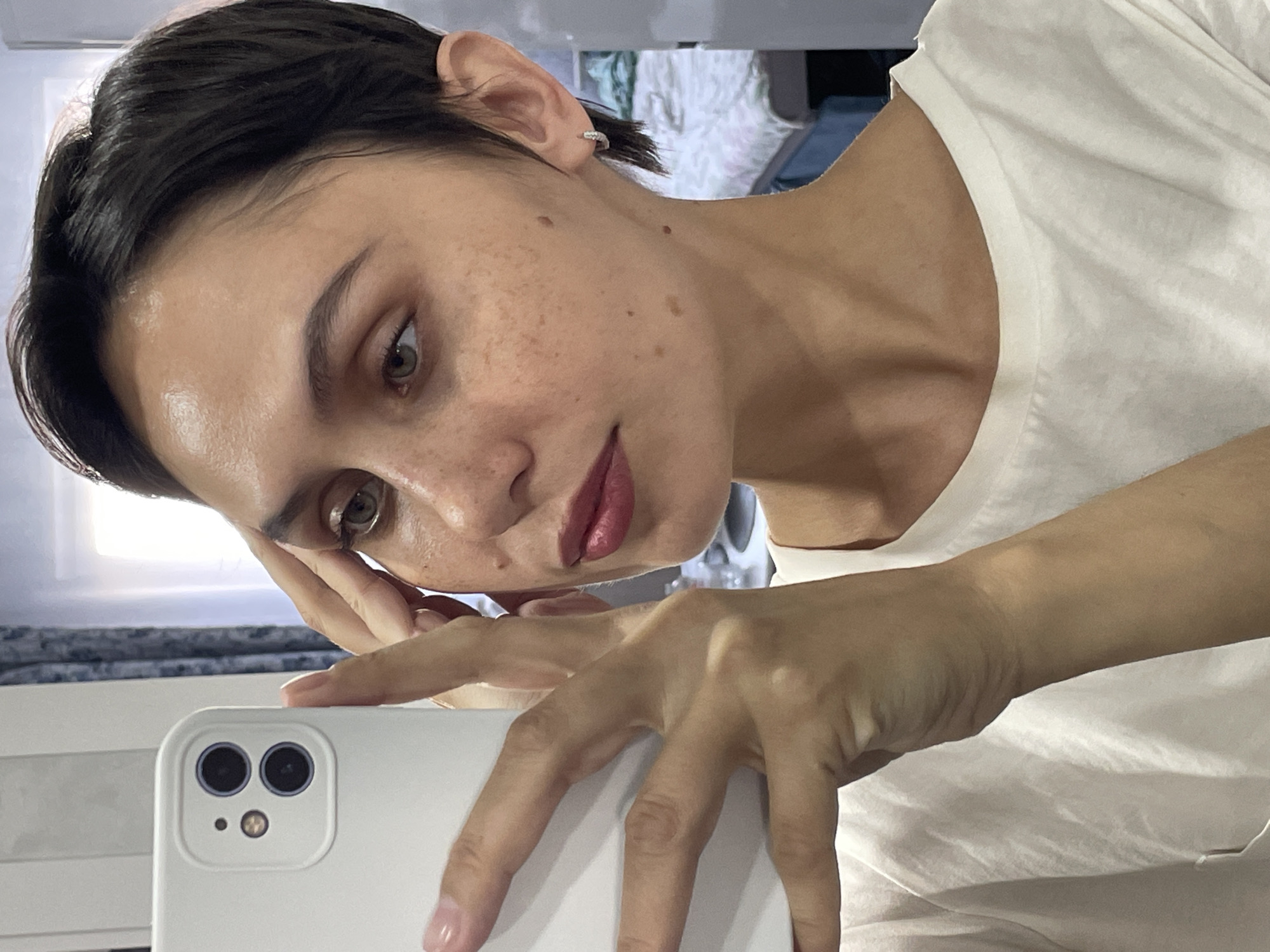 A woman with short dark hair is taking a selfie with a white smartphone. She is wearing a white T-shirt. Her head is tilted, and she is looking at the phone screen. Her left hand is on her face, and her right hand is holding the phone. The background is blurred, but a window and part of a room can be seen. It is unclear if the location is Izhevsk, Russia, as there is no readily identifiable information in the image to confirm this.