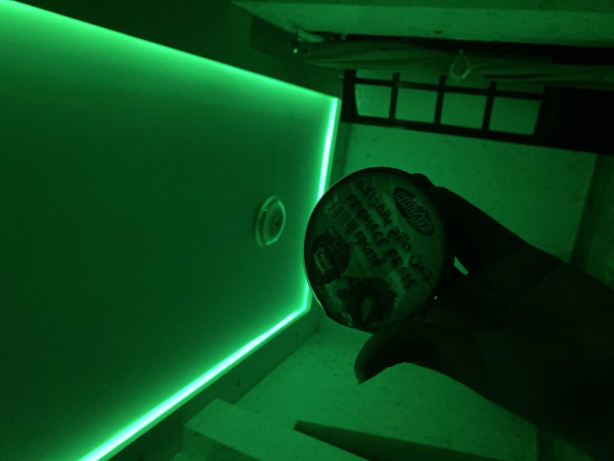 The image depicts a dimly lit interior space, possibly a room, illuminated by prominent green LED strip lighting along the ceiling's edges. The camera angle is from below, looking upwards. In the foreground, a hand is visible holding a round container, likely yogurt or a similar dairy product. Text is visible on the container, though partially obscured and difficult to read due to the lighting and angle. The text appears to be in French and possibly Arabic, with some words like "FRAISE" (strawberry) and "YAOURT" (yogurt) discernible. In the background, a tall, dark structure resembling a ladder or shelving unit is present. The overall atmosphere is dark and atmospheric, with the green light casting a strong, uniform hue over the entire scene. No specific indicators place this scene definitively in Baraki, Algeria, beyond the provided prompt context.