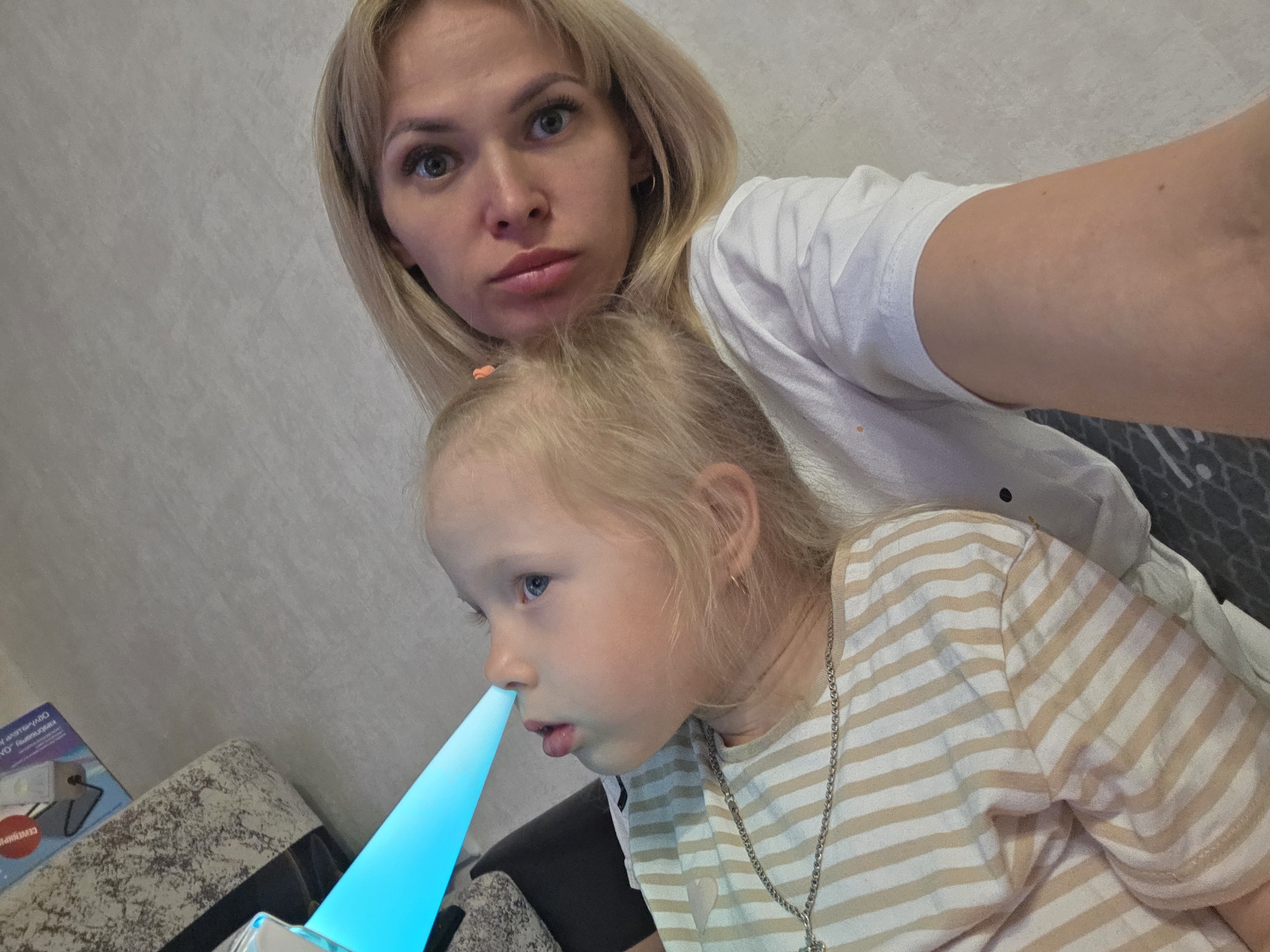 A woman and a child are in what appears to be a residence. The woman, with blonde hair and fair skin, is wearing a white t-shirt. The child, who also has blonde hair and fair skin, is wearing a striped t-shirt and a necklace. A bright blue light is directed at the child's nose, seemingly originating from a device. The background suggests a room interior with light-colored walls and a box on a table. The image may have been taken in Chaikovsky, Russia.