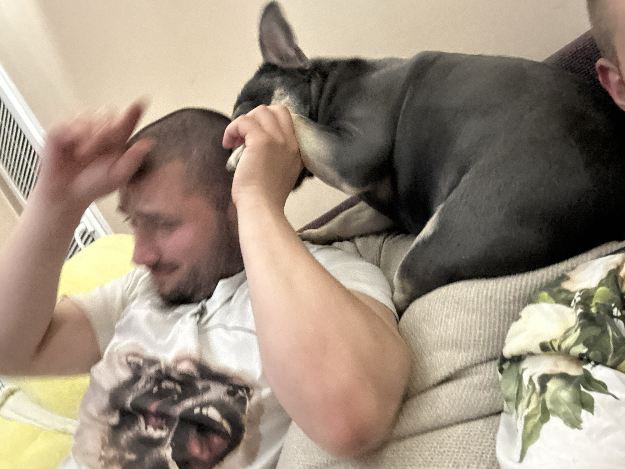 A person is lying down on a couch, with a small black and tan dog resting on top of them. The person's head is turned to the side, with their eyes closed, and their arm is raised, with their hand near their face. The dog is looking towards the person's face, with its head tilted to the side. The scene appears to be indoors, possibly in a living room. The person is wearing a white t-shirt with a graphic on it, and the dog is dark-colored with tan markings. The couch is upholstered in a neutral fabric, and there is a decorative pillow with a floral pattern visible in the foreground. The overall lighting is dim, suggesting it may be evening or night.
