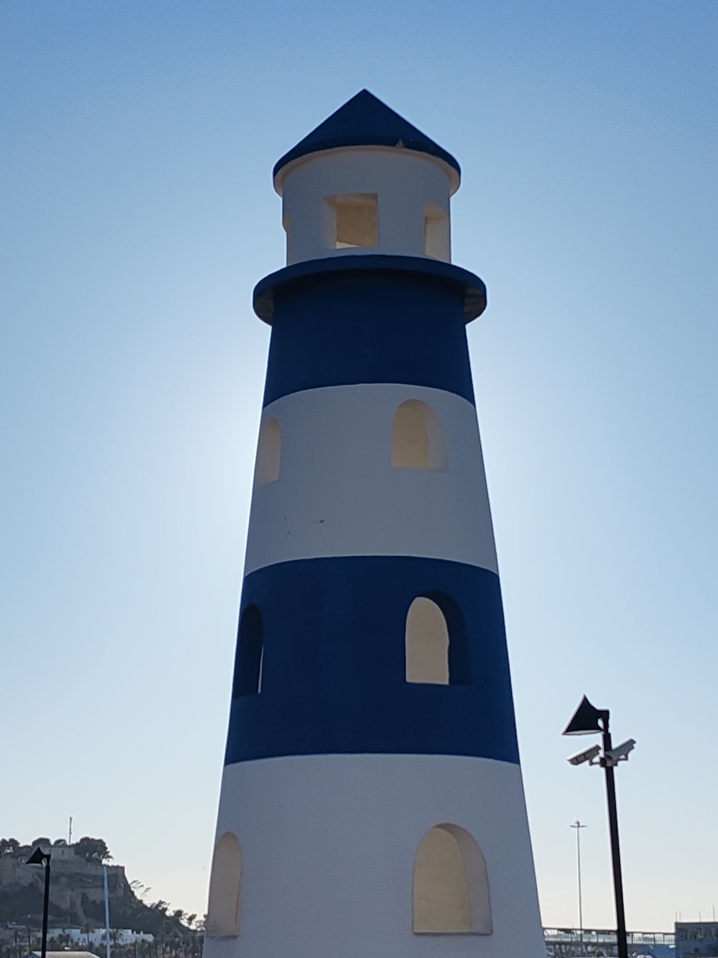 The image shows a decorative blue and white lighthouse in Dénia, Spain. It's a sunny day with a clear blue sky. The lighthouse has a traditional striped pattern with arched window openings. The structure is located near the harbor area, with a streetlight in the foreground and buildings along the coast in the background. The image captures the coastal atmosphere of Dénia, a popular tourist destination.