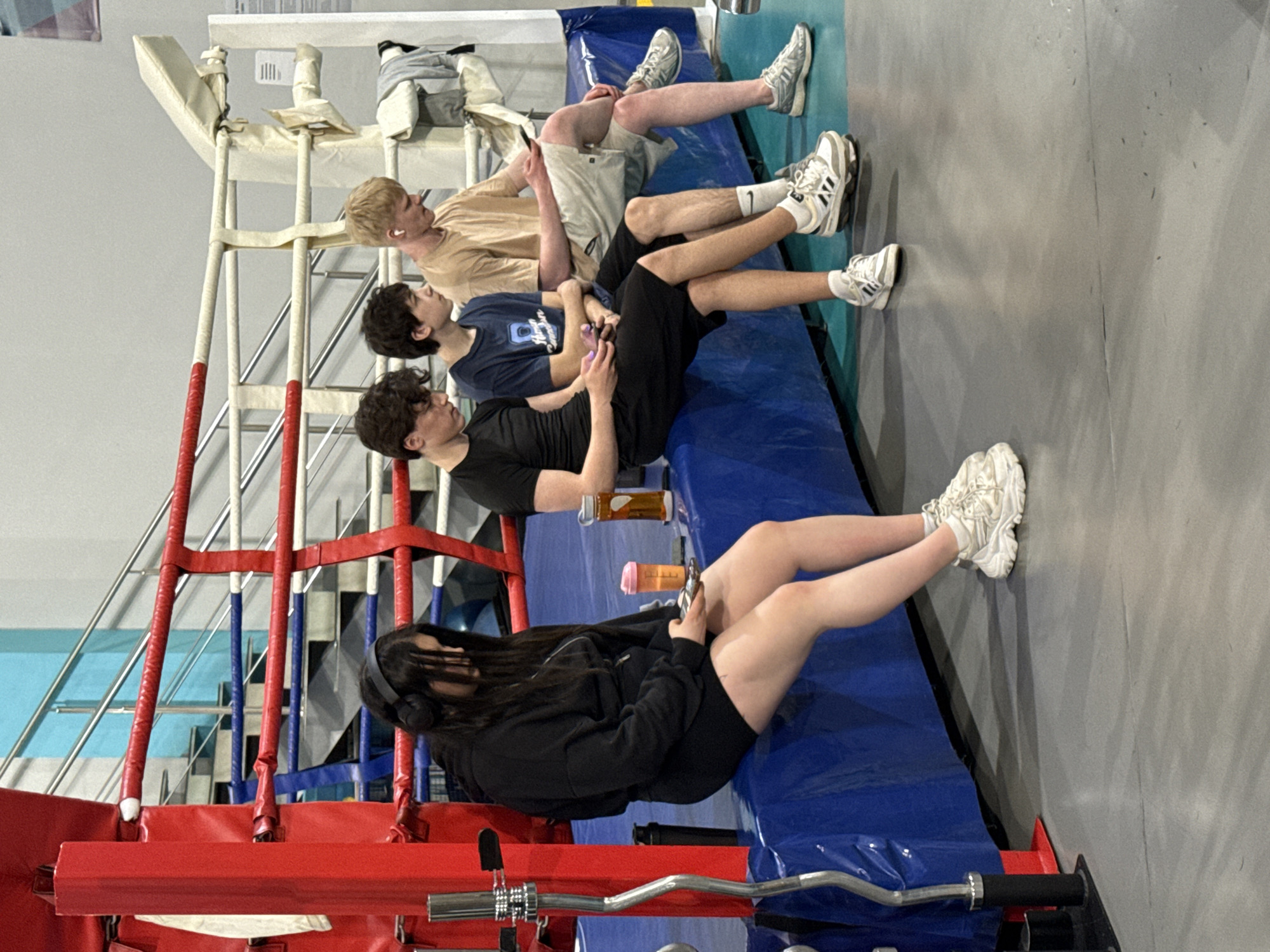 Four people are sitting on the padded blue border of a boxing ring. From top to bottom, the first person is wearing a tan shirt and khaki shorts. The second person is wearing a blue t-shirt and black shorts. The third person is wearing a black t-shirt and black shorts. The fourth person is wearing a black sweatshirt and black shorts. The boxing ring has red, white, and blue ropes. A barbell is visible in the foreground. The location is indoors. The city is not discernible from this image.
