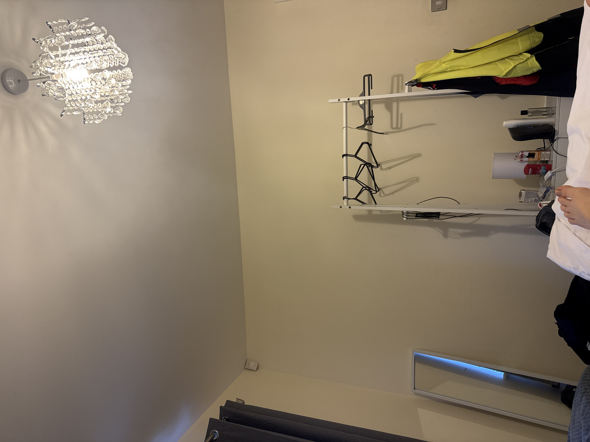 The image shows an interior wall featuring a crystal chandelier, a two-tiered shelf, and a mirror. The chandelier, with a silver base, is affixed to the left wall. On the opposite wall, a white metal shelf holds various items, including clothes, containers, and a small lamp. A mirror with a white frame is positioned near the bottom of the wall. The location is Swindon, United Kingdom.