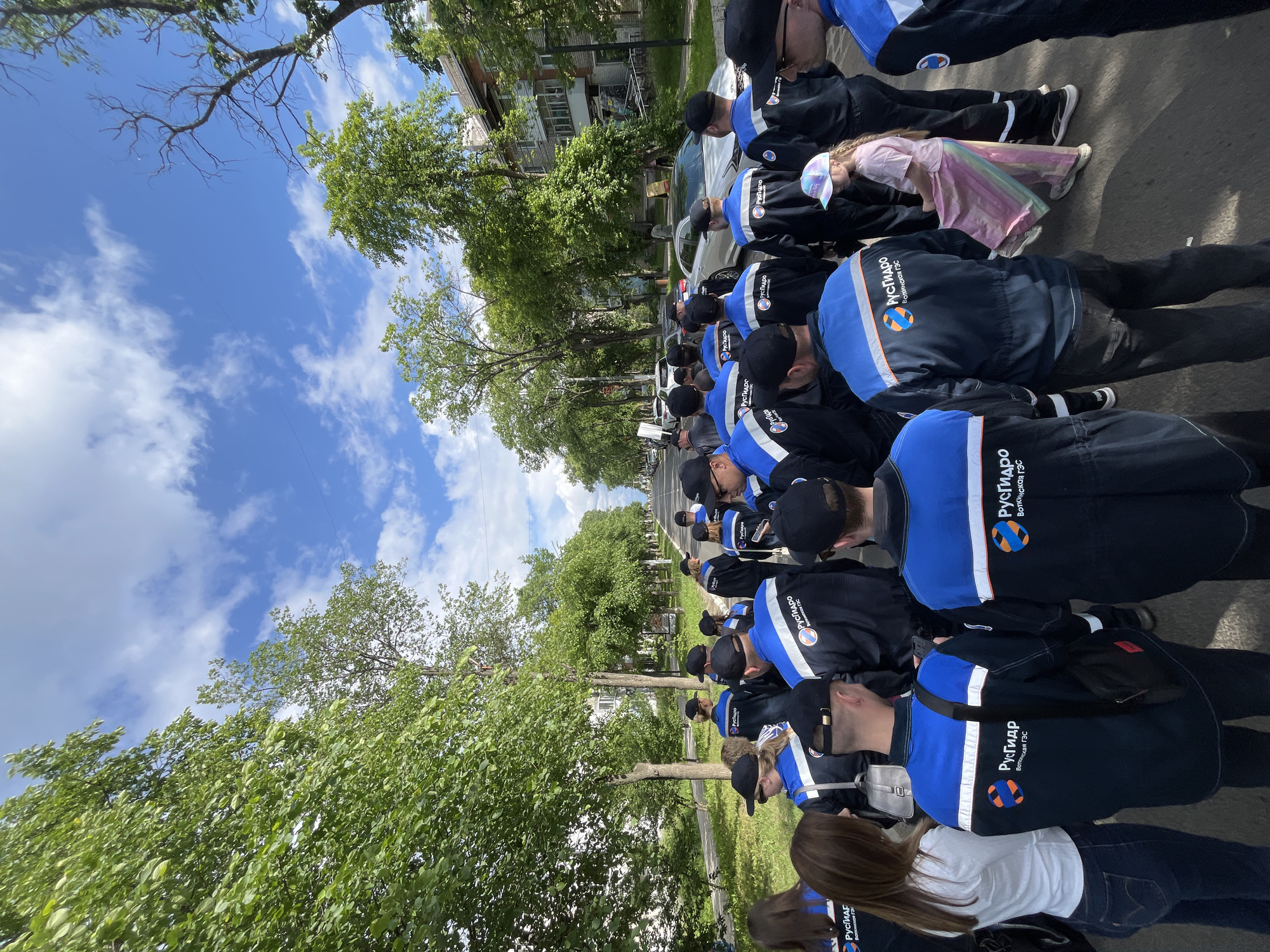 A group of approximately 20 people, including men, women, and one young girl, are standing and walking on a paved road lined with trees and residential buildings. Most of the adults are wearing matching dark blue jackets with orange and white stripes on the shoulders and reflective white stripes on the sleeves, and some are wearing black caps. The jackets have a logo with Cyrillic text that appears to read "РусГидро" and "Воткинская ГЭС". The young girl is wearing a pink t-shirt and a colorful, iridescent skirt and a patterned baseball cap. In the background, there are cars parked and driving on the road, and a multi-story brick building. The sky is blue with scattered white clouds. The overall impression is a group outing or event in an urban or suburban setting in Chaikovsky, Russia, possibly related to the Votkinsk Hydroelectric Power Station.