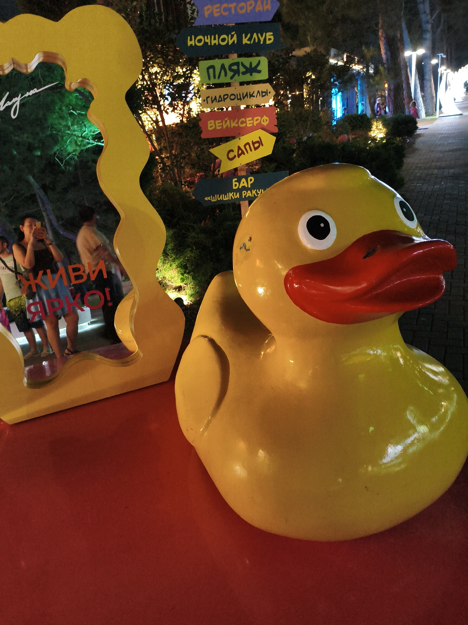 This photograph captures a bright yellow, oversized rubber duck sculpture positioned prominently in the foreground. The duck has large, black-and-white eyes and a red beak. Behind the duck, a large, bright yellow abstract sculpture curves upwards and to the left, reflecting some of the surrounding environment. In the background, several directional signs with Russian text are visible, indicating various locations such as "RESTORAN," "НОЧНОЙ КЛУБ" (Night Club), "ПЛЯЖ" (Beach), "ГИДРОЦИКЛЫ" (Jet Skis), "ВЕЙКСЕРФ" (Wakeboard), "САНЫ" (Sleds), "БАР" (Bar), and "«ШИШКИ РАКУШКИ»" (Pinecones Seashells). The scene appears to be an outdoor recreational area, possibly a promenade or park, at night. The ground is a deep red surface, and a paved walkway with lampposts is visible further in the background. A few people are partially visible in the mid-ground, with one woman taking a photo with her phone. The overall setting suggests a lively atmosphere, likely a tourist destination in Gelendzhik, Russia, known for its beachfront attractions.