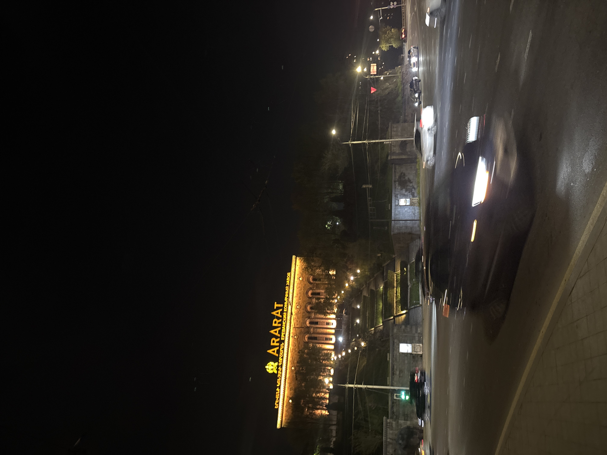The image shows a nighttime scene in Yerevan, Armenia. The "ARARAT" sign in yellow lettering is visible on a building. The building is likely the Yerevan Brandy Company, as the sign includes the phrase "ԵՐԵՎԱՆԻ ԿՈՆՅԱԿԻ ԳՈՐԾԱՐԱՆ" (Yerevani Konyaki Gortsaran), which translates to "Yerevan Brandy Factory." Cars are seen driving on a road adjacent to the building. The environment is dark, with the scene illuminated by streetlights and car headlights. There's a road and some vegetation visible.