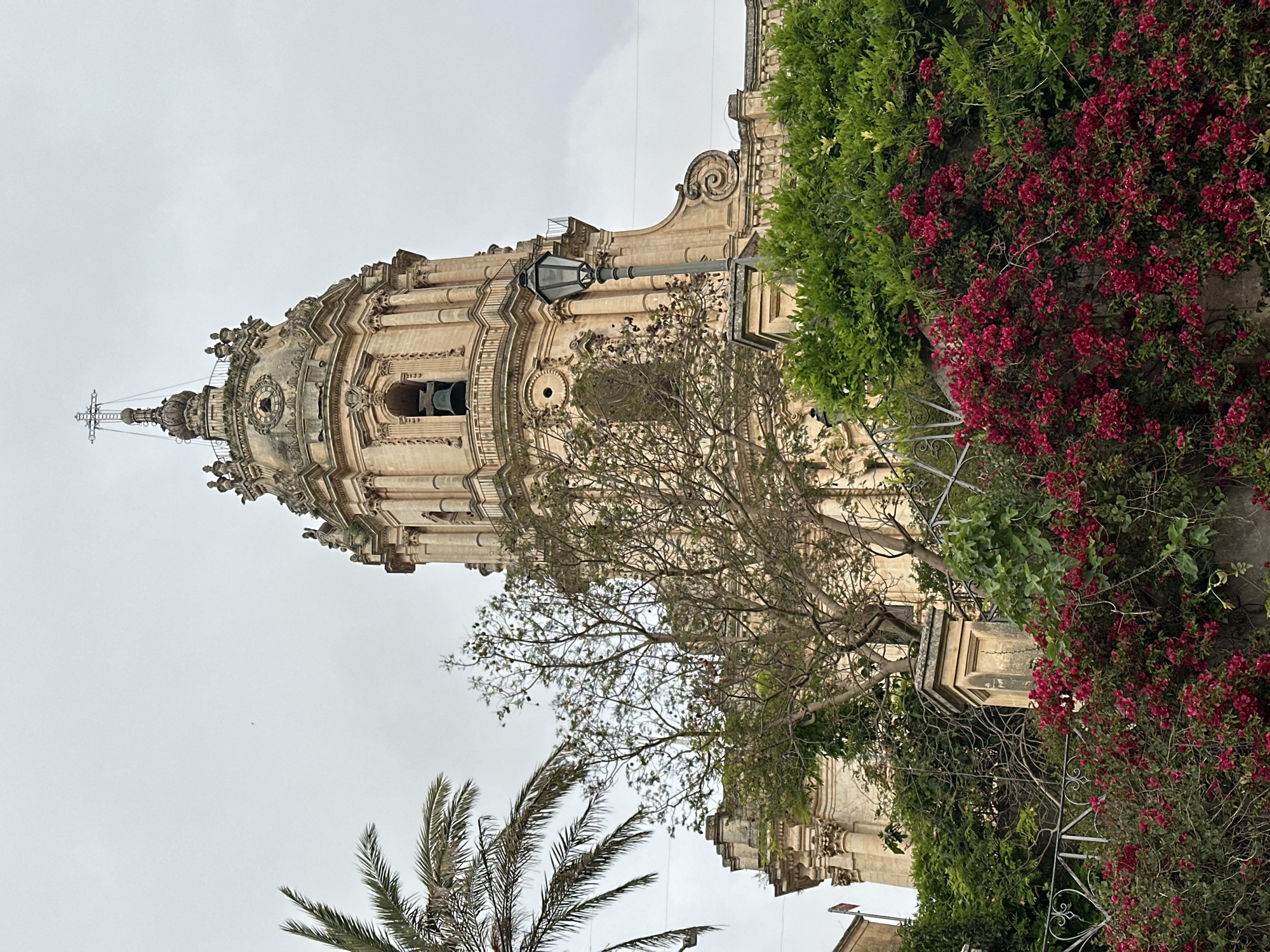 The image shows the dome of the Saint George Cathedral in Modica, Italy, partially obscured by trees and flowering bushes. The dome is of ornate Baroque design, made of light-colored stone, with a clock visible near the top. A lamppost extends from the facade of the building. The sky is overcast.