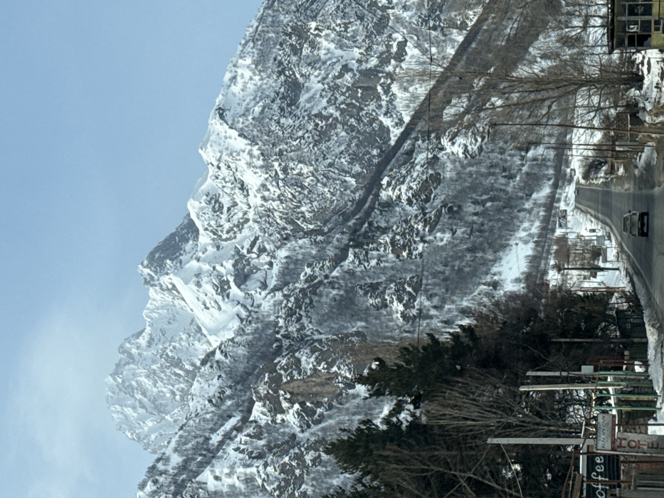 The image depicts a snow-covered mountain landscape within the designated city of Goristsikhe, Georgia, under a clear, light blue sky. A two-lane asphalt road, partially cleared of snow, extends from the foreground into the distance, leading towards a mountain pass.
On the left side of the road, several low-rise structures are visible, including one with prominent signage displaying "Coffee" horizontally and "HOTEL" vertically in English, along with what appears to be Georgian script. Tall, bare deciduous trees and some snow-laden coniferous trees line this side, interspersed with multiple utility poles and their associated lines.
A light-colored sedan is observed traveling away from the viewer in the middle of the road. Further in the distance along the road, additional vehicles and indistinct structures can be faintly discerned. The right side of the road also features bare trees, utility poles, and a small, unidentifiable structure in the foreground.
The background is dominated by massive, jagged mountain peaks and slopes, extensively covered in white snow. Darker rock faces and sparse, snow-dusted vegetation are visible on the mountainsides. The scene indicates a cold, clear winter day. No individuals are overtly identifiable.