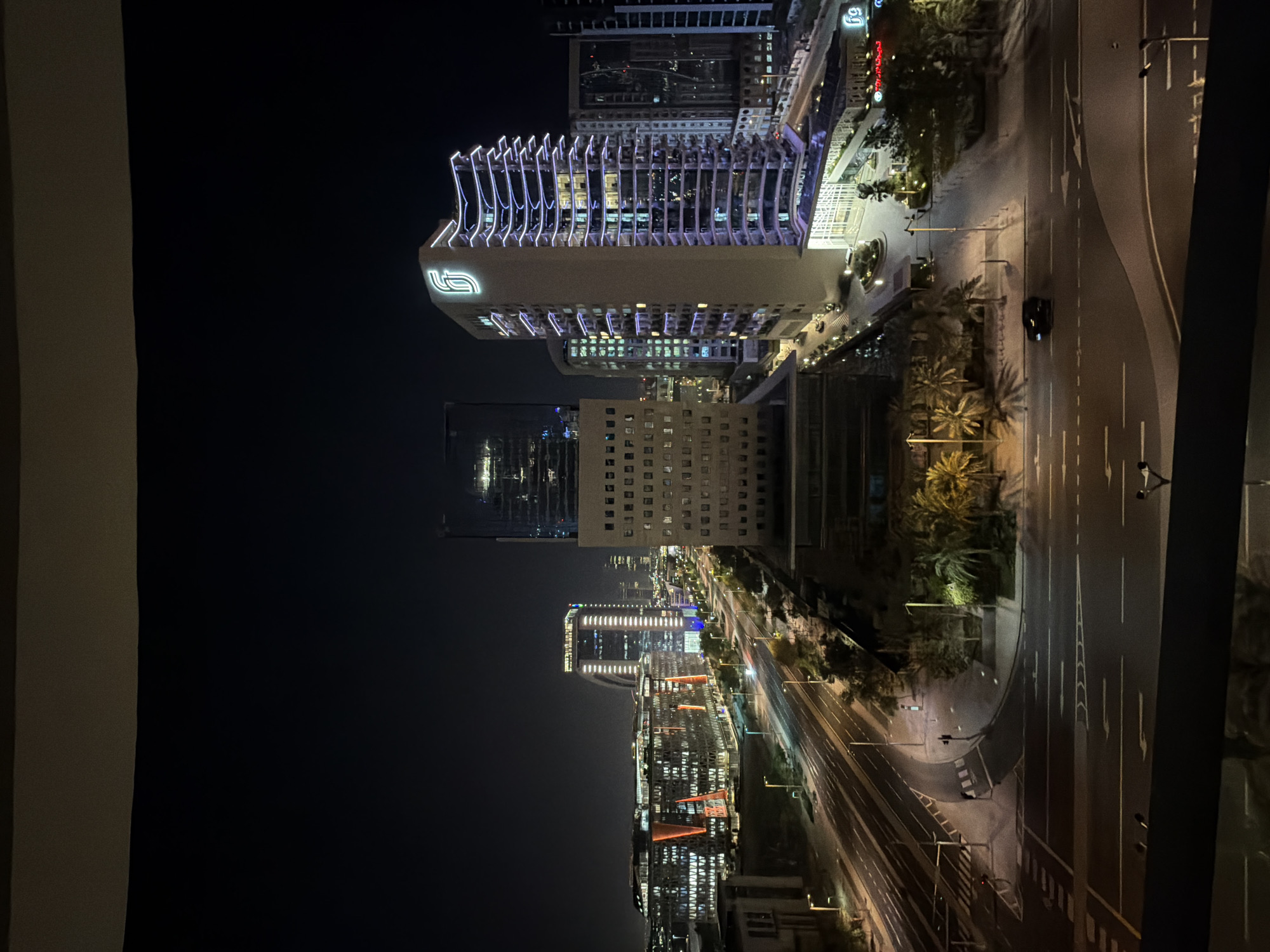 A nighttime aerial view captures a modern cityscape characterized by illuminated buildings and a well-lit road network. In the foreground, a dark, multi-lane road curves through the scene, with white lane markings and directional arrows visible. A single black car is driving on the road. The middle ground features a distinct tall, cream-colored building with a prominent logo on its upper section, outlined by a purple, wave-like light design along its facade. Adjacent to it are other buildings, some with intricate glass facades and orange and white lighting. Palm trees are scattered throughout the landscape, their shadows cast on the ground. In the background, more buildings of varying heights and designs are illuminated against the dark sky, contributing to a vibrant urban panorama. The image appears to be taken from a high vantage point, possibly a balcony or window, looking out over the city.