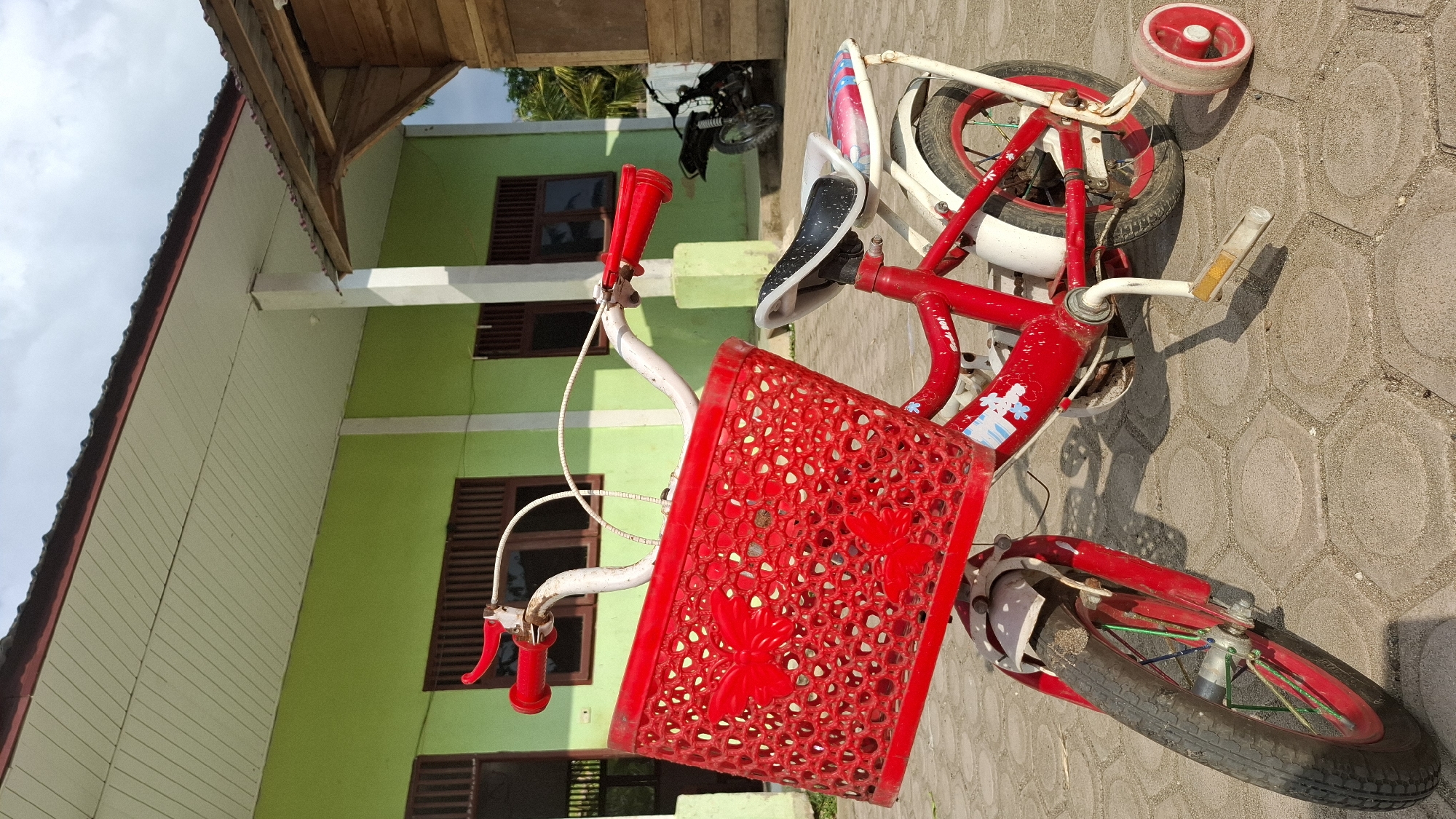 A bright red child's bicycle with training wheels is parked on a light-colored hexagonal paver patio in front of a light green building with a brown roof. The bicycle has a red basket adorned with butterfly cutouts on the handlebars. The building appears to be a school or community center, with multiple windows visible. To the right of the building, a portion of a wooden structure is visible. A black motorcycle is parked in the background. The sky is cloudy.