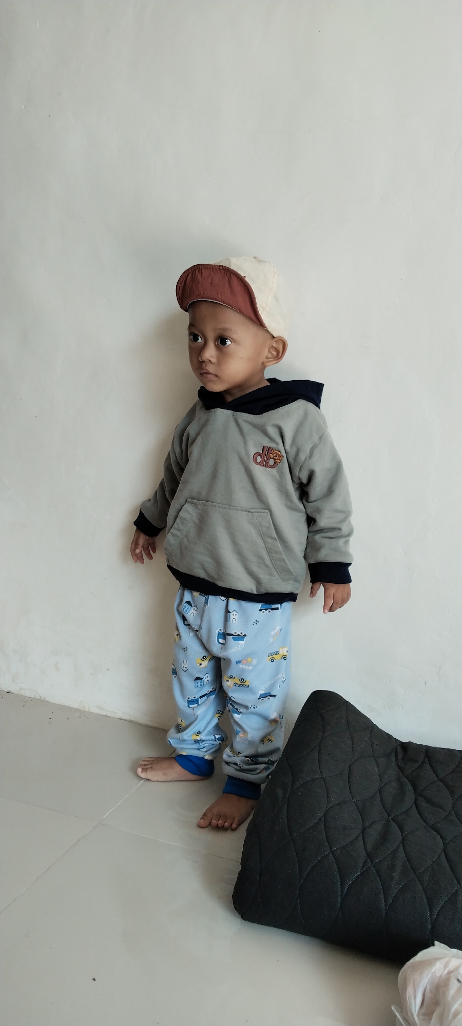 A young child, estimated to be between 2 and 4 years old, stands facing left against a textured white wall. The child is wearing a grey hooded sweatshirt with a dark navy hood lining and blue cuffs, blue sweatpants adorned with a cartoon print of construction vehicles and houses, and a two-toned baseball cap with a brown brim and a cream-colored crown. The child's feet are bare. To the right of the child, a dark, quilted rectangular object, possibly a cushion or mat, rests on a light-colored tiled floor. The lighting appears to be natural and ambient. The setting suggests an indoor environment, likely a residence, in Kecamatan Pati, Indonesia.