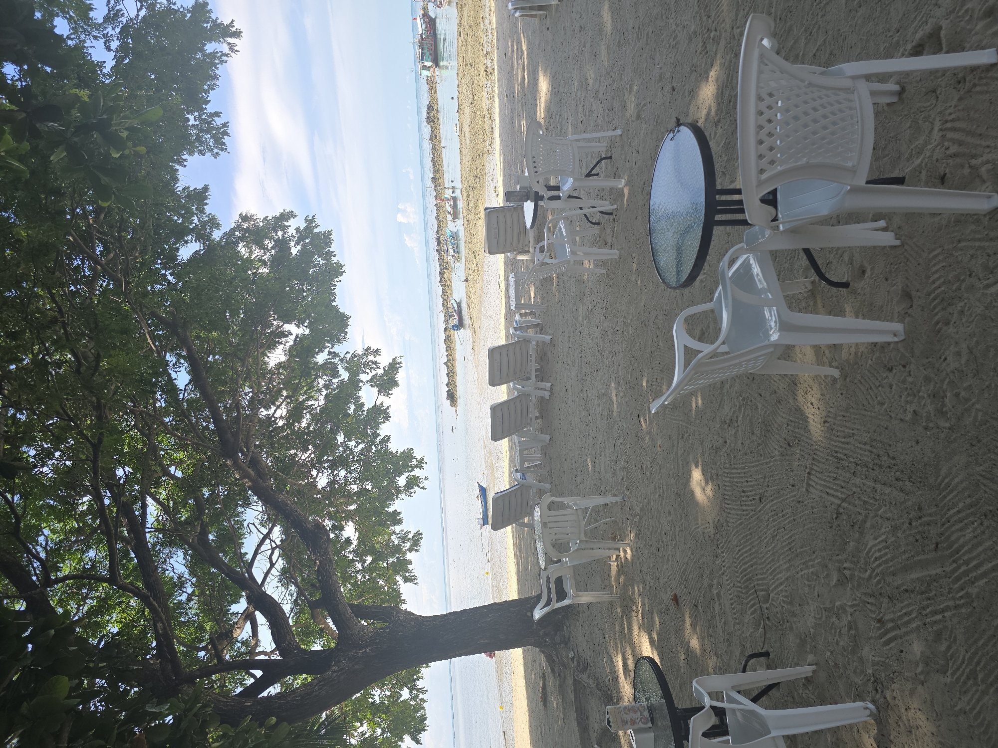 The image depicts a serene beach scene in Baan Chaweng Noi, Thailand. In the foreground, several white plastic chairs are arranged around round tables on the sandy beach. Some of these tables have glass tops, reflecting the sunlight. A large tree with dense green foliage extends from the left side of the frame, casting shadows on the sand.
Beyond the seating area, the beach slopes gently towards the water. A line of empty lounge chairs is visible in the middle ground, suggesting a quiet moment at this establishment. The shallow water is calm and clear, with a few small fishing boats anchored offshore. The horizon is dotted with more boats and a distant island or landmass. The sky above is a pale blue with scattered white clouds, indicating fair weather. The overall impression is one of a peaceful and inviting seaside cafe or rest area.