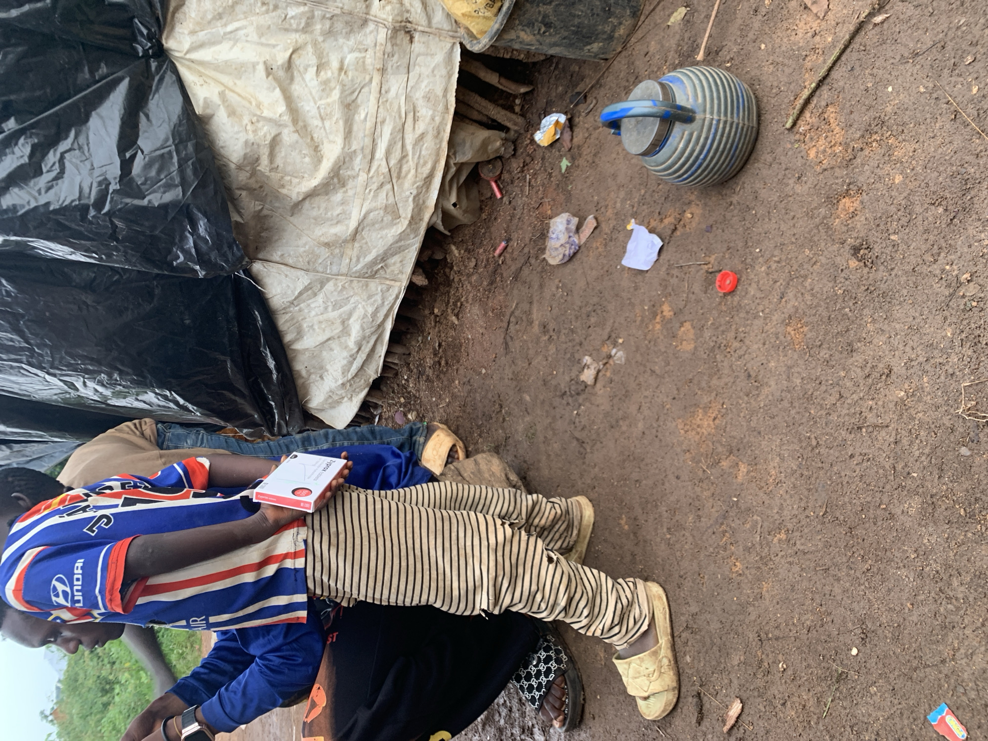 This image depicts a group of people in a makeshift shelter constructed from a black tarp and a white, weathered material. The ground is composed of damp, reddish-brown soil with scattered debris. Two individuals are prominently featured, seated on the ground. One person wears a blue and white striped long-sleeved shirt with red accents and the Hyundai logo on the sleeve. This person is holding a white and red box of medication with visible text. They are also wearing striped pants. Another person is visible behind them, wearing blue jeans.
In the background, a grey and blue ridged watering can is resting on the ground, next to scattered pieces of paper and what appear to be small red cylindrical objects. The overall scene suggests a humble dwelling or temporary shelter, possibly in a rural area given the natural ground and materials used for construction. The lighting suggests it is daytime. The image captures a moment of rest or interaction within this environment. The text on the medication box is partially visible and reads "Ziprolac capsule".
