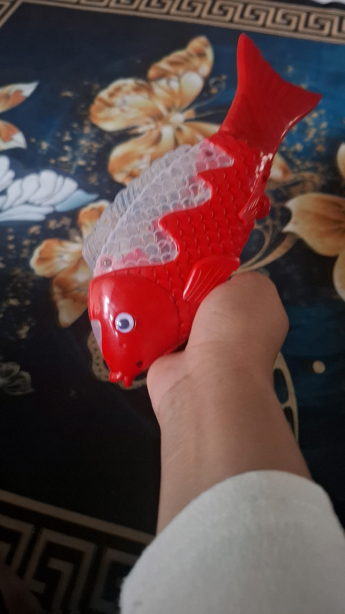 A hand is holding a red and white toy fish against a dark blue background with a floral pattern. The toy fish has a translucent white fin on its back and a red body with a white eye. The background also features a decorative border with a geometric pattern along the top and bottom. The scene is indoor, possibly a home environment. No specific location within Bandar Lampung, Indonesia can be determined from the image.