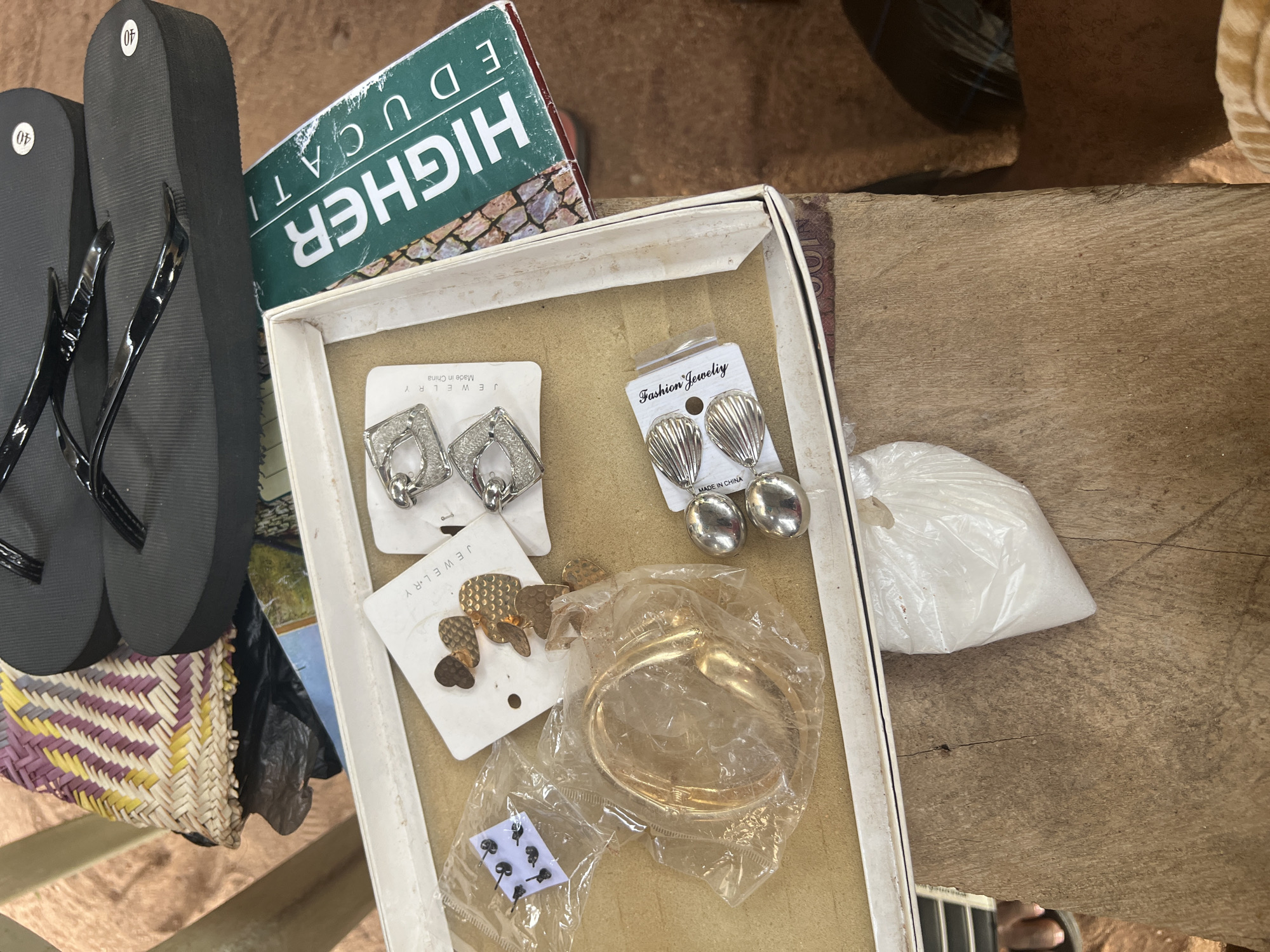 The image shows a close-up, overhead view of a collection of items on a wooden surface, likely in Jalingo, Nigeria. In the background, a pair of black flip-flops with shiny straps rests on a woven mat. To the left of the flip-flops, a portion of a black plastic bag is visible. A white cardboard box is open in the foreground, containing various pieces of jewelry. These include a pair of silver-toned earrings with a square, open-work design, and another pair of silver-toned earrings shaped like scallops. There is also a bracelet or bangle, encased in plastic, which appears to be gold-toned. Next to the bracelet is a small, clear plastic bag containing several pairs of small, black stud earrings. To the right of the box, a green book with white text that reads "HIGHER EDUCAT" is partially visible. Beneath the box, a white plastic bag filled with white granular material, possibly sugar, is present. The wooden surface has visible grain and some cracks. The lighting suggests an indoor or shaded outdoor setting.