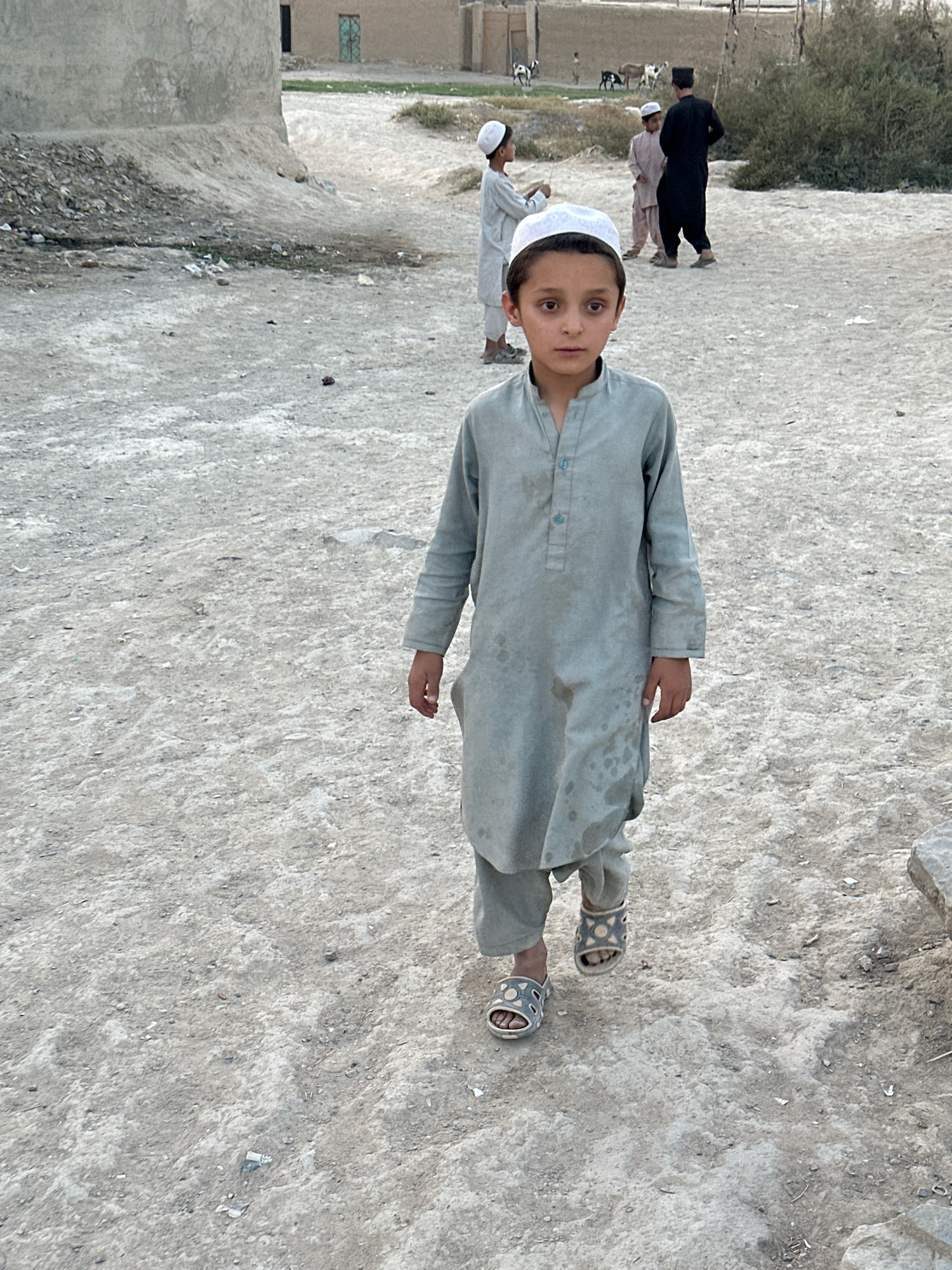 The image depicts a young boy wearing a light blue kurta and trousers, and a white cap, walking on a dirt path in Kuchlak, Pakistan. He is facing the camera, with a neutral expression. In the background, other individuals are visible, including another boy in a similar outfit, and a man in a black kurta and trousers. There are also a few goats grazing in the distance. The environment appears to be an outdoor setting with dusty ground, some buildings, and sparse vegetation. The lighting suggests it is daytime.