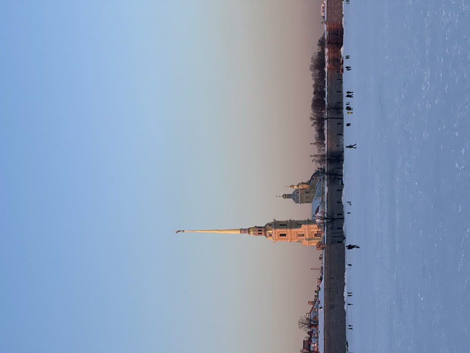 An outdoor scene in Saint Petersburg, Russia, depicts the Peter and Paul Fortress and Cathedral from across the frozen Neva River during winter. The sky is clear, transitioning from light blue overhead to a pale orange-pink near the horizon, indicative of a low sun angle.
In the foreground, a vast expanse of snow-covered ice, likely the frozen Neva River, occupies the lower half of the frame. Scattered across this icy surface are approximately two dozen human figures, appearing small due to distance, engaged in walking or similar activities.
Midground features the robust, light-colored stone walls of the Peter and Paul Fortress, running horizontally across the image. Patches of snow are visible on the wall tops, interspersed with bare deciduous trees.
Dominating the background skyline is the Peter and Paul Cathedral, situated within the fortress. Its tall, slender golden spire, topped with a distinct weathervane figure, reflects the warm, low sunlight, as do its associated golden domes and the main structure of the cathedral. Additional smaller buildings and domes of the fortress complex are visible behind the primary wall. The scene is illuminated by natural light, casting long shadows.