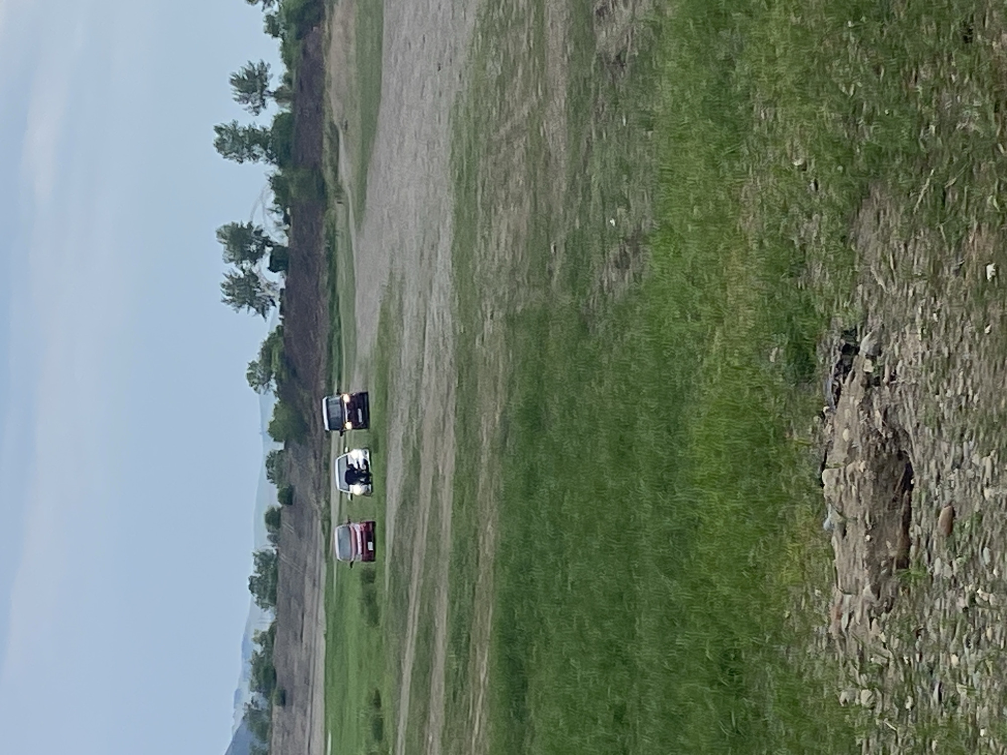 The image depicts an outdoor scene with several vehicles on a grassy and dirt terrain. Three cars are visible: a red car on the left, a white car in the middle, and a dark-colored van or SUV on the right. The white car has a person partially visible inside. In the background, there are hills and trees, with a cloudy sky overhead. The environment suggests a rural or semi-rural location, possibly a park or an undeveloped area. No specific landmarks or details indicate the city of Chernogorsk, Russia. The overall impression is a casual outdoor gathering or stop with vehicles.