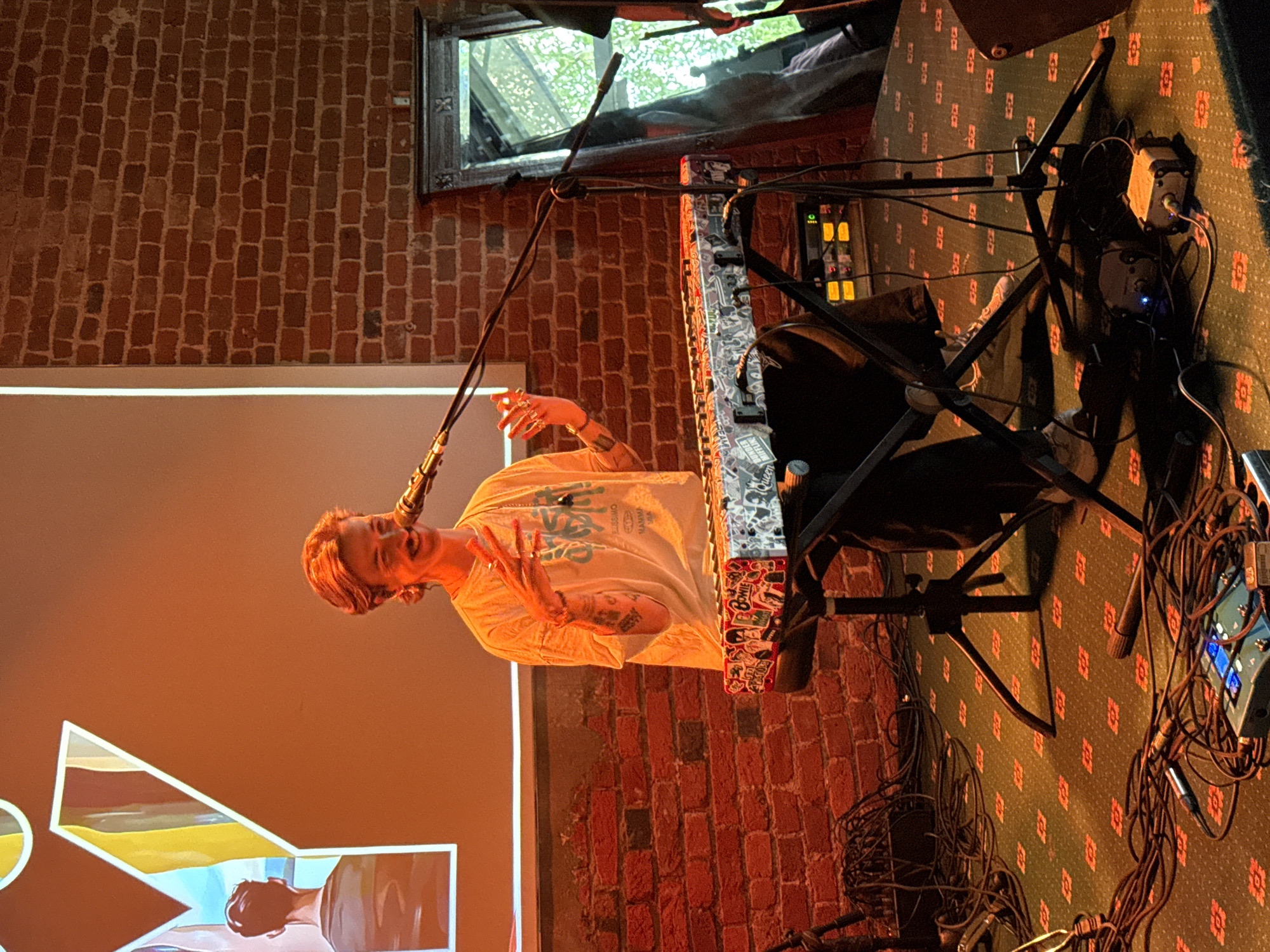 A musician performs on stage in what appears to be a brick-walled venue. The performer, a fair-skinned individual with short, light hair, is positioned behind a keyboard stand. They are wearing a light-colored t-shirt and dark pants. Their left arm is extended with an open hand gesture, and the microphone is in front of them.
The keyboard has various stickers. Cables and electronic equipment clutter the floor near the keyboard stand. A window is visible in the background, above the musician's head.
A large, illuminated logo is on a screen next to the performer. The venue has a patterned carpet. Brickwork can be seen on the walls.
Given the Cyrillic alphabet in the image on the screen, it's reasonable to infer the location is Moscow, Russia.