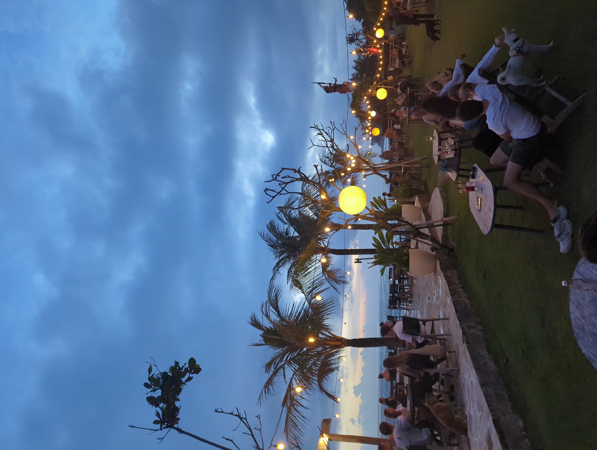 The image captures an outdoor evening scene in Mengwi, Indonesia. The sky is overcast with dark blue clouds. String lights are strung among palm trees and bare trees, illuminating the area. People are seated at tables on a grassy area next to a stone pathway. The ocean is visible in the background. A performer stands on a pole in the distance.