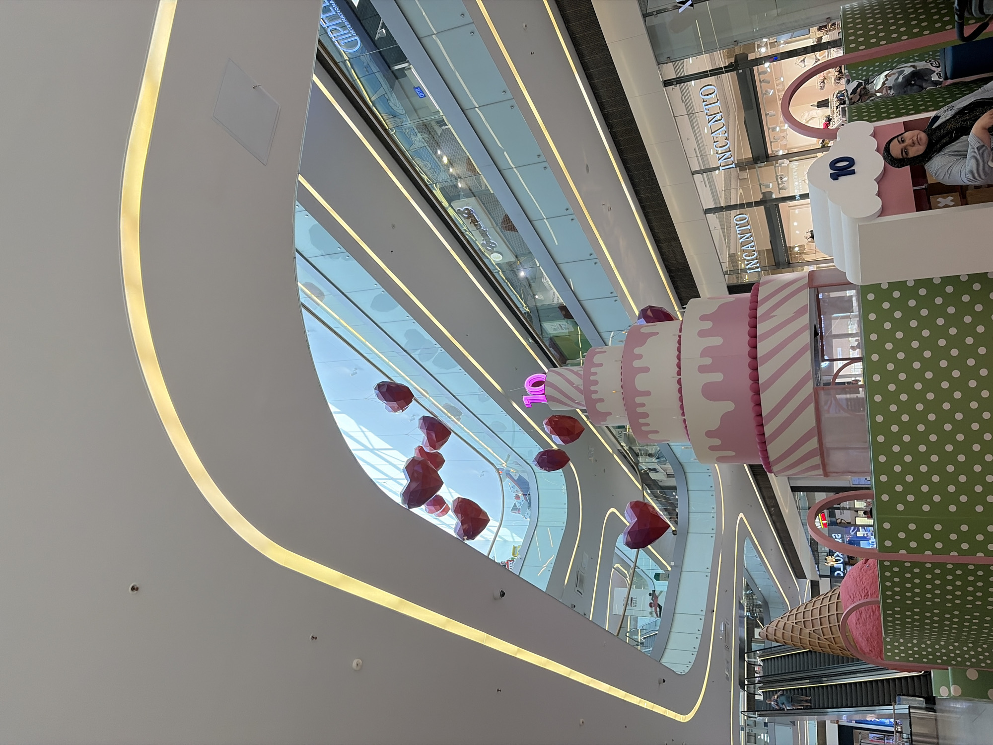 This media file shows an interior view of a modern shopping mall in Saint Petersburg, Russia. The scene is dominated by the mall's architecture, featuring curved white walls with integrated yellow lighting. Several escalators are visible, indicating multiple levels within the mall. In the foreground and mid-ground, there are large decorative elements resembling a cake and an ice cream cone, suggesting a festive or promotional event. The cake is multi-tiered with pink and white frosting and pink stripes. The ice cream cone is oversized and pink. Scattered throughout the upper part of the image are several red, heart-shaped balloons, adding to the celebratory atmosphere.
In the background, through glass panels, stores with signs are visible. The word "INCINTO" is clearly legible on multiple store facades. A woman wearing a headscarf is partially visible on the right side of the image, looking towards the camera. Other people are seen on the escalators in the distance, appearing as small figures.
The lighting suggests it is daytime, and the indoor setting implies no specific weather conditions are relevant. The overall mood conveyed by the decorations and lighting is cheerful and commercial. The presence of "INCINTO" provides a specific location cue, likely identifying a brand or store within the mall.