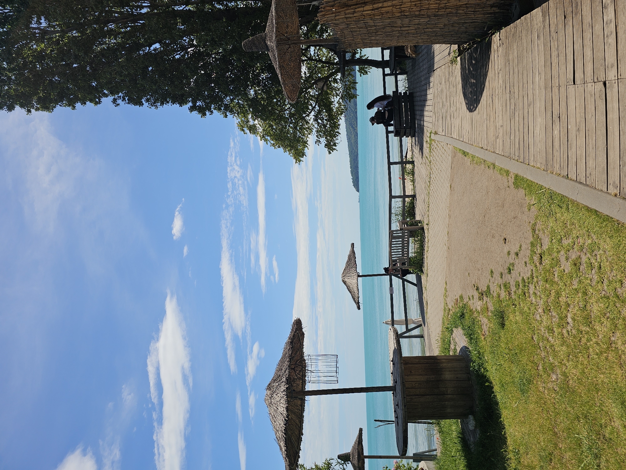 The image depicts a waterfront scene with a boardwalk and beach umbrellas. The camera is tilted, so the sky appears on the left and the ground on the right. The sky is blue with scattered white clouds. The water is turquoise and stretches to the horizon. Along the boardwalk, there are benches and people sitting. Straw beach umbrellas are positioned along the water's edge. The ground is covered with grass and some paved areas.