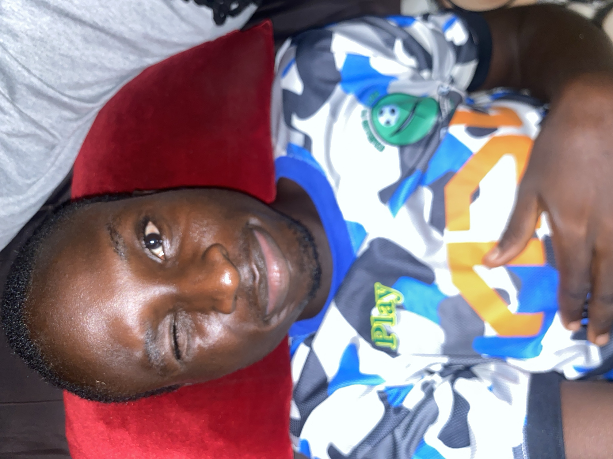A medium shot of a Black man, likely lying down, possibly on a red pillow. He has one eye open and a slight smile on his face. He is wearing a colorful jersey with a camouflage pattern and the word "Play" on it. The image does not provide any location details that confirm it is in Barden Yana, Nigeria, so this can't be confirmed.