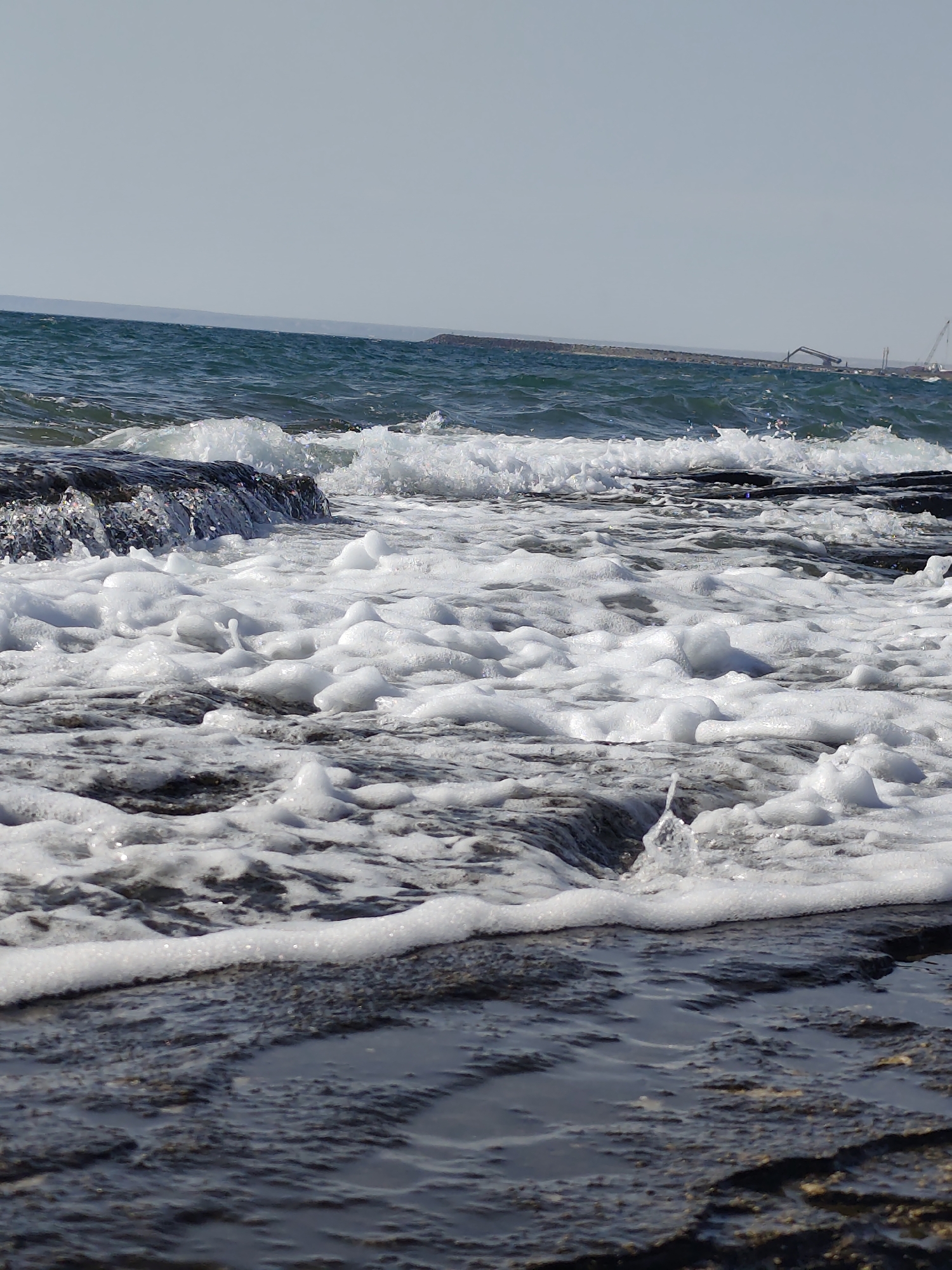 A low-angle, medium shot captures a churning sea with foamy white waves crashing against dark, wet rocks in the foreground. The water is a deep teal with lighter, frothy areas where the waves break. The sky above is a pale, clear blue. In the far distance, a long, dark breakwater stretches across the horizon, with industrial cranes and structures visible on it. The image focuses on the dynamic interaction of the water and the rocky shore, with no people or animals present. The scene evokes a sense of a natural coastal environment, possibly near a port or industrial area.