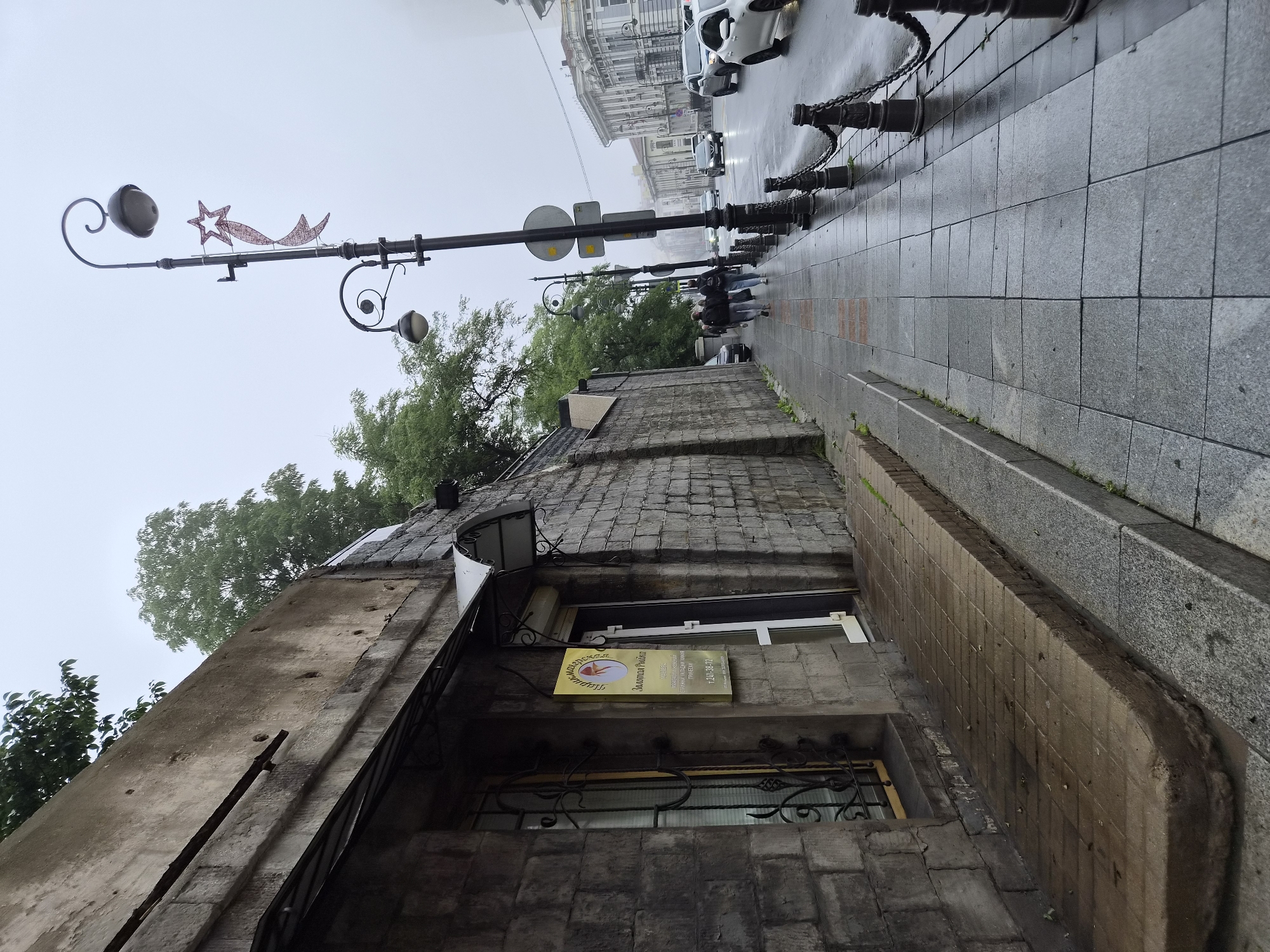 This image captures a street scene in Vladivostok, Russia, on an overcast day. The perspective is from the sidewalk, looking down a street lined with buildings and trees.
On the left side of the frame, a textured stone wall forms the side of a building. Above the wall, green foliage is visible, suggesting trees or bushes. An awning extends from the building, and a yellow sign with Cyrillic lettering and an image of a woman's head is mounted near an entrance. The sign appears to advertise a beauty salon or similar establishment, as indicated by text that includes "Золотая Рыбка" (Golden Fish). A window with decorative black metalwork is also present. The sidewalk in front of the building is paved with square tiles, and a raised, curved concrete pathway leads to the entrance.
To the right of the sidewalk, a black metal railing with chains separates the pedestrian area from the street. Several lampposts with decorative elements, including a red star and ribbon decoration on one, are spaced along the sidewalk. A few people are walking away from the camera down the sidewalk.
The street itself is wet, indicating recent rain or a damp environment. Several cars are driving on the street, including a white sedan on the right and a silver SUV. In the background, more buildings with classic architectural details are visible, and a hazy or foggy atmosphere contributes to the muted lighting of the scene. The overall impression is of a slightly melancholic, urban environment on a day with poor weather.