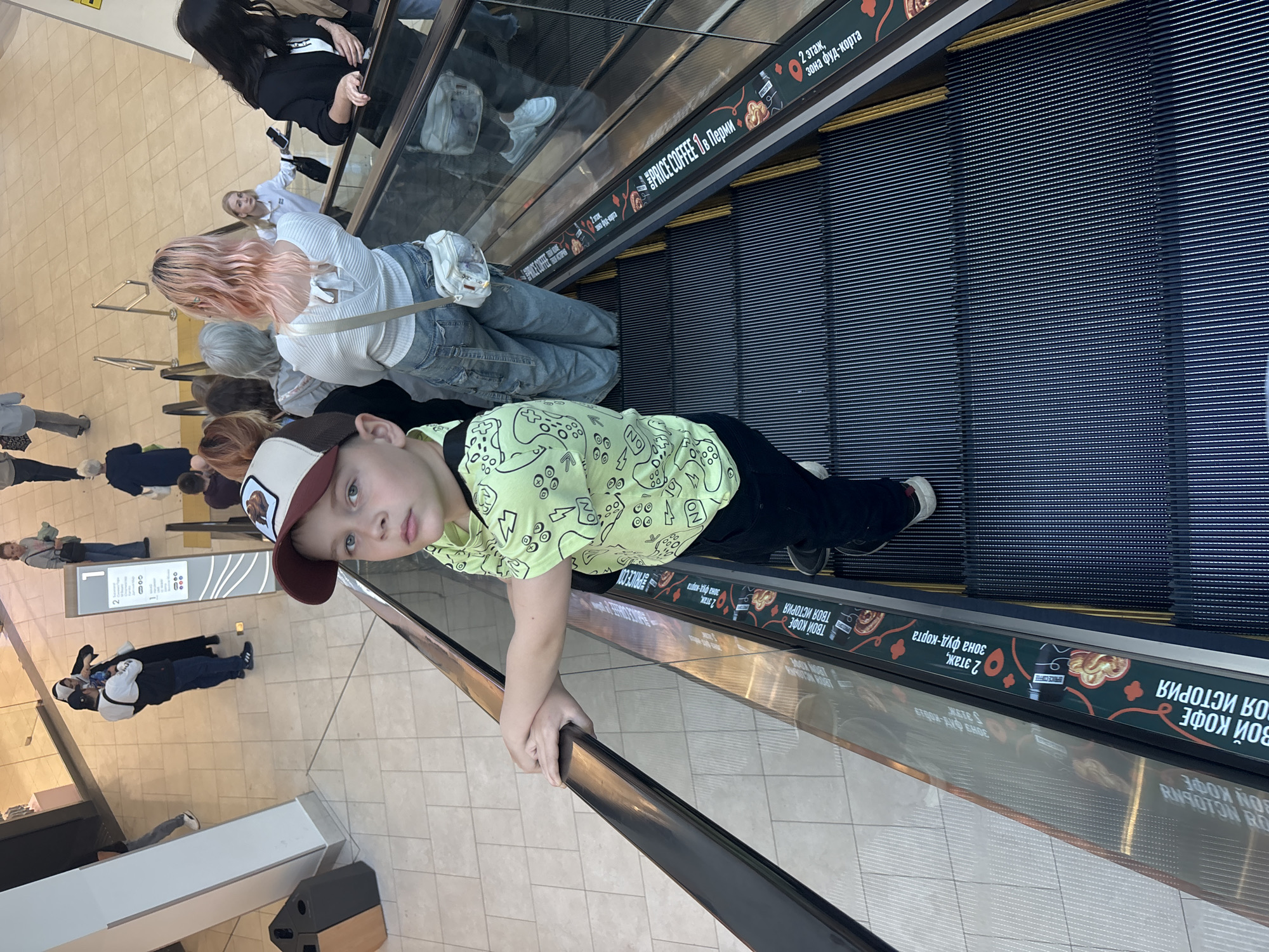 The image depicts a high-angle view of a young boy on an ascending escalator inside a multi-story commercial building, likely a shopping mall, in Perm, Russia.
In the foreground, a boy approximately 8-10 years old stands on the escalator, facing the camera. He has light brown hair, blue eyes, and a neutral expression. He wears a beige baseball cap with a dark red brim and a brown graphic, a bright lime green short-sleeved t-shirt with black game controller-themed graphics (including "ON" and "NO" text), and black pants. His hands are clasped over the black rubber handrail of the escalator. The escalator steps are dark with yellow demarcation lines along the edges, and the side panel is clear glass.
To the boy's right, an informational strip runs along the base of the glass panel, displaying text in Russian: "PRICE COFFEE В Перми" (PRICE COFFEE in Perm), "ТВОЙ КОФЕ ТВОЯ ИСТОРИЯ" (YOUR COFFEE YOUR STORY), and "2 этаж, зона фуд-корта" (2nd floor, food court zone), along with graphics of coffee, pretzels, and map pins. This confirms the location as Perm.
Behind the boy on the escalator, several individuals are also ascending. A woman with wavy, light pink hair, wearing a white long-sleeved top with a V-neck back and light blue jeans, carries a white crossbody bag. Further back, an older woman with short grey hair is visible.
To the right of the ascending escalator, an adjacent escalator is descending. A woman with dark hair, wearing a black blazer over a white top, holds a mobile phone. Another woman in a white long-sleeved shirt is visible further down the descent.
On the lower floor, visible in the background, numerous people are walking on a light-colored tiled floor. A tall, rectangular off-white pillar stands on the left. Beside it, a grey information sign displays a large "1" at the top, likely indicating the ground floor. Near the pillar, a man in a black and white jacket and dark pants interacts with a woman wearing a black hijab and a long dark dress. The man holds a blue bottle. A two-part waste receptacle (brown and black) is situated near the pillar. Other individuals are scattered across the lower level, moving through the space. Glass walls and shopfronts are reflected in the background.