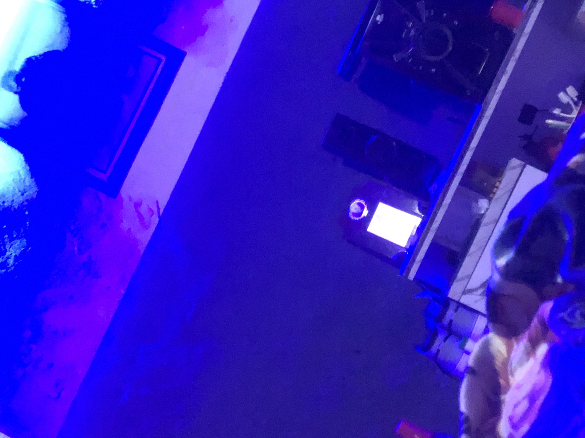 The image depicts an indoor environment in the city of Unknown, Unknown, characterized by predominant artificial blue and purple lighting. The perspective is tilted and upward-angled.
In the immediate foreground, blurred masses of textile, exhibiting hues of pink, purple, and blue, are visible on the left and center. A dark, reflective, curved object, possibly an item of footwear, is partially discernible on the bottom right. A rounded, light-toned form, consistent with a human limb, is partially visible on the bottom left.
A white, horizontal surface, appearing to be a shelf or cabinet top, spans the mid-ground. Positioned on this surface, from left to right: an electronic device with a central glowing white rectangular display and a circular, illuminated blue element on its top; a dark, rectangular electronic device; and a larger, dark, rectangular electronic device, likely audio equipment, featuring circular structural elements. A red plastic cup rests on the far right of this white surface.
The upper portion of the image displays a wall illuminated intensely with blue and purple light. A dark-framed, rectangular object, possibly an artwork, is mounted on the upper-right wall; its contents are obscured by the lighting. No discernible human faces are present in the image.
