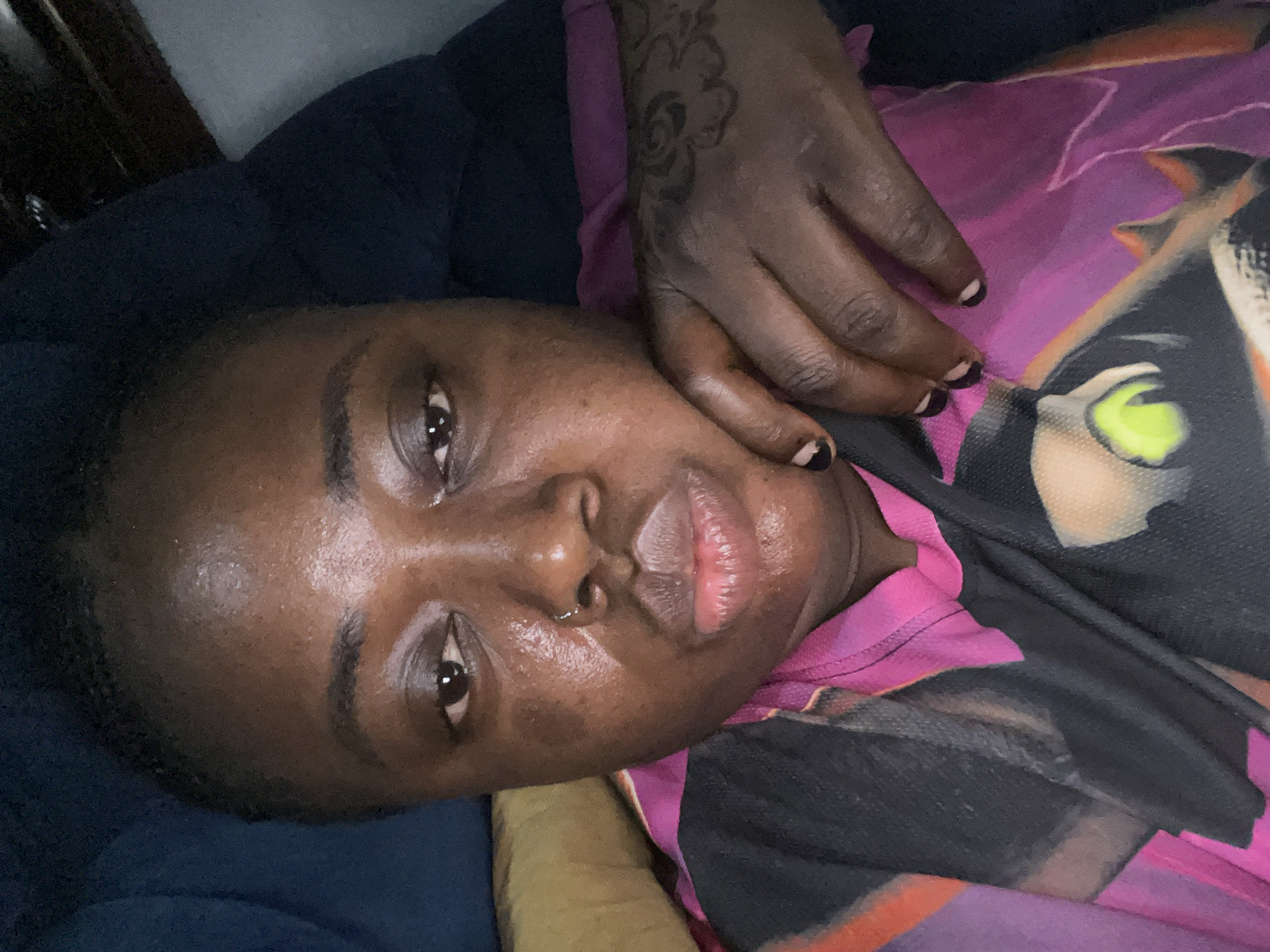A person with dark skin lies on a blue surface. Their right hand, adorned with a flower-shaped tattoo, rests on their face. The person wears a pink and black patterned garment. A gold-colored object is visible underneath the person's head. There is no information to support the person being in Kubwa, Nigeria.